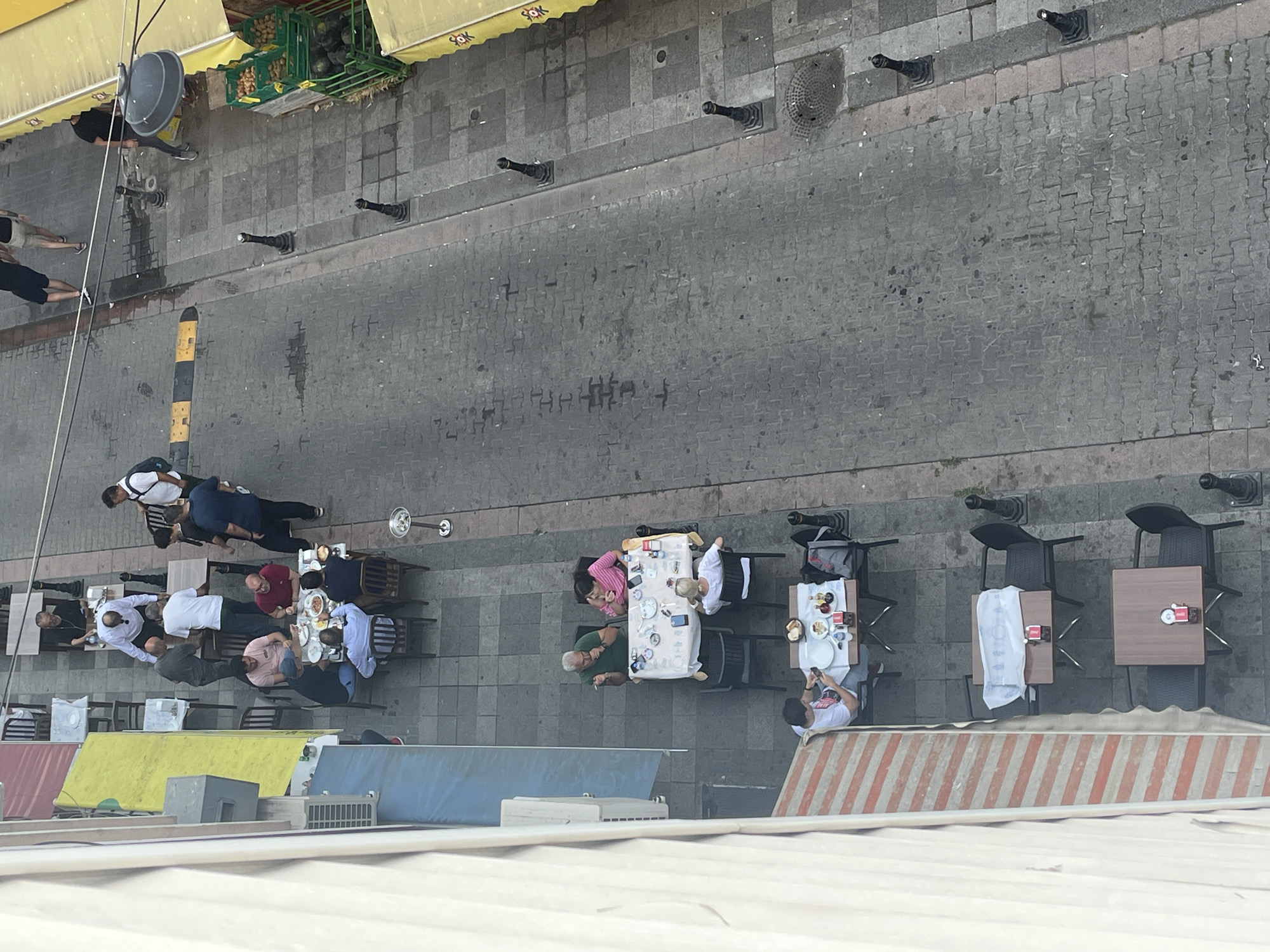 An aerial, high-angle, wide shot captures a street scene in Kadıköy, Turkey, with outdoor seating for a restaurant or cafe on the left and a cobblestone street on the right. Several tables with patrons are visible. On the left, a person in a white shirt sits at a table covered with a white tablecloth, looking at their phone. Next to them, another table has a white cloth with food and drinks, and three people are seated. One person in a green shirt is smoking, another in a pink striped shirt is eating, and a third person in a white shirt is seated. Further up the street on the left, several tables are occupied by people eating and talking. On the right side of the street, there is a yellow awning with the word "ŞOK" visible, and green crates containing produce, possibly kiwis and watermelons. Pedestrians are visible walking on the street. The overall atmosphere appears to be casual and social, with people enjoying a meal outdoors.