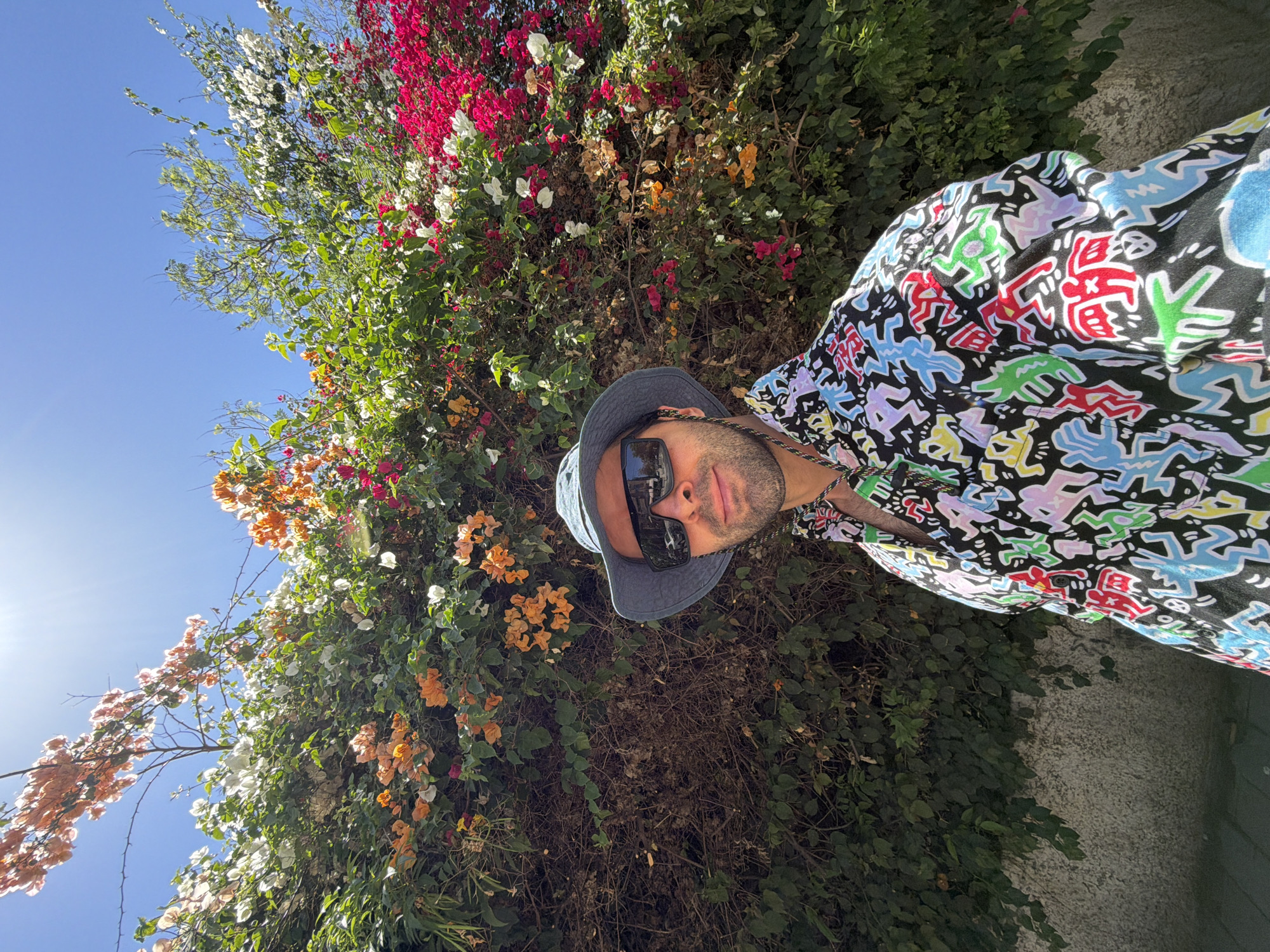 A man is depicted in a close-up, outdoor portrait shot in Tel-Aviv, Israel. He is positioned slightly off-center to the right, facing the camera with a subtle smile. He wears a faded blue bucket hat and large, dark, reflective sunglasses that obscure his eyes, with faint reflections of an urban environment visible on their surface. A multicolored lanyard is visible around his neck, and he has short facial hair. His shirt is black, featuring a vibrant, repeating pattern of multicolored stylized figures in red, blue, green, yellow, white, and light purple. Behind him is a dense, tall hedge predominantly composed of flowering bougainvillea in shades of fuchsia, white, and orange. The foliage ranges from lush green to drier brown patches. A bright blue sky with a strong sun glare in the upper left corner forms the upper background. A textured light-colored wall, partially covered by green vegetation, is visible in the lower left background.