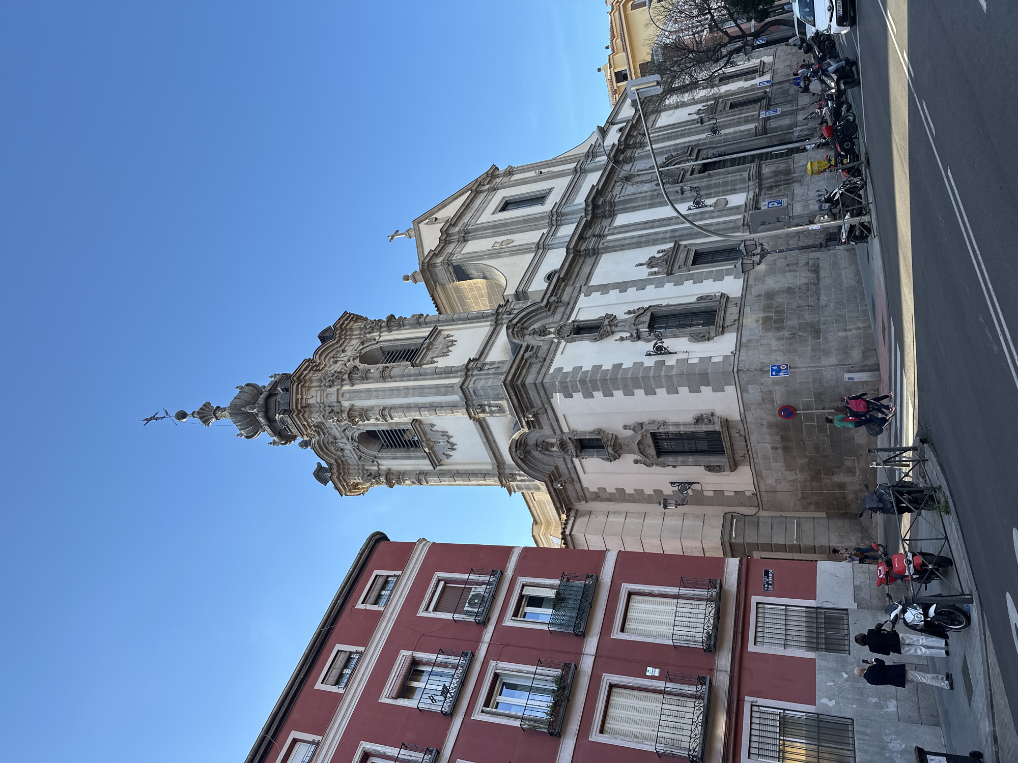 The image depicts a street scene in Madrid, Spain, dominated by the ornate Baroque facade of the Pontifical Basilica of St. Michael. The basilica, with its white stone and intricate detailing, towers over a red building with wrought-iron balconies on the left.
People are visible on the sidewalk in front of the buildings, including pedestrians and individuals standing near parked motorcycles. The street itself is paved and features double yellow lines dividing traffic lanes. Several motorcycles and scooters are parked along the sidewalk. Street signs and parking indicators are mounted on the building’s exterior. The environment is bright, suggesting a sunny day.