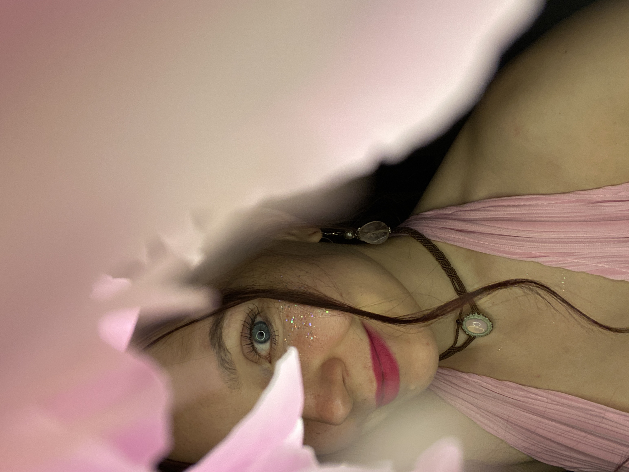 The image presents an angled, close-up portrait of a young woman, partially framed by blurred, soft pink elements resembling petals or flowing fabric in the foreground. She is lying down, her face turned upwards, gazing directly into the camera with piercing blue eyes. Her dark hair cascades around her, with strands visible across her face and chest.
She is adorned with bright pink lipstick and a scattering of iridescent glitter on her left cheek and under her eye. Her attire includes a light pink, pleated garment, visible across her shoulder and chest. A dark brown, braided necklace is prominent, featuring two distinct pendants: a clear, faceted, crystalline bead near her neck, and a smaller, round, opalescent pendant with a light-colored inlay lower on her chest.
The background is dark and out of focus, enhancing the subject's prominence. The overall aesthetic is dreamy and artistic, suggesting a staged photoshoot. The woman's direct gaze conveys a sense of engagement and a captivating allure. No specific architectural or environmental location cues are discernible within the image itself; however, the context provided states the location is Chaikovsky, Russia. There is no visible text in the image.
