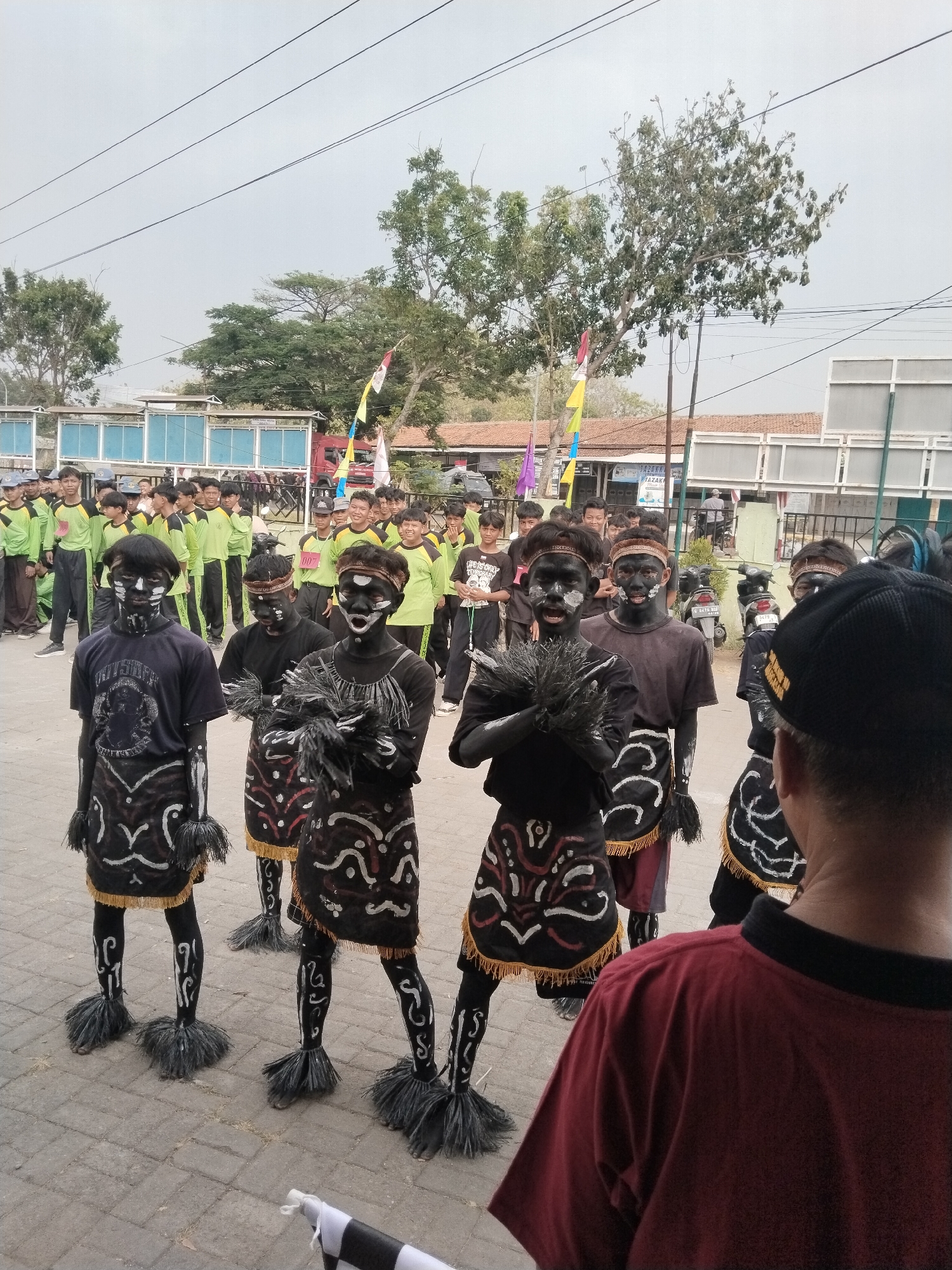 The image shows a group of young men in traditional attire, their faces and bodies painted black with white accents. They are standing on a paved street in what appears to be a public event. Some of the men are holding decorative fluffy objects in their hands. They are wearing patterned skirts and leggings, with feathered attachments on their feet. Behind them, a larger group of young men in green and yellow uniforms are visible, along with some adults. In the background, there are buildings, trees, and electrical wires. The scene suggests a cultural performance or parade taking place in an outdoor urban setting.
