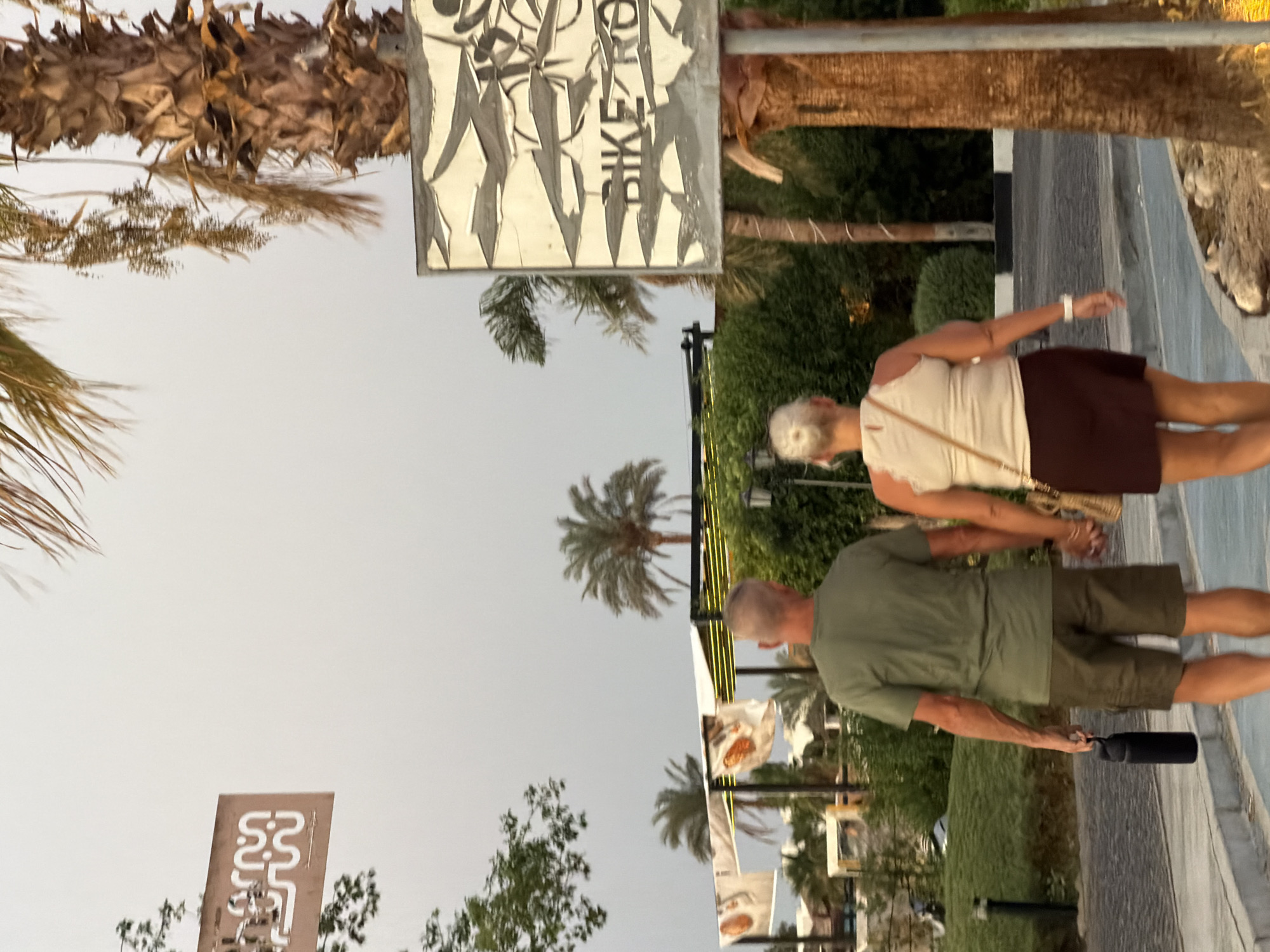 A couple, a man and a woman, walk away from the camera hand-in-hand on a paved walkway. The man, wearing a green t-shirt and olive shorts, carries a black water bottle in his left hand. The woman, wearing a cream-colored tank top and a maroon skirt, has a tan crossbody bag. They are walking past a damaged sign that reads "BIKE RO". In the background, palm trees and lush green foliage are visible. Buildings with white canopies are also present, along with draped fabric banners. The sky is overcast and appears to be either early morning or late evening. This scene is located in Sharm El Sheikh, Egypt.