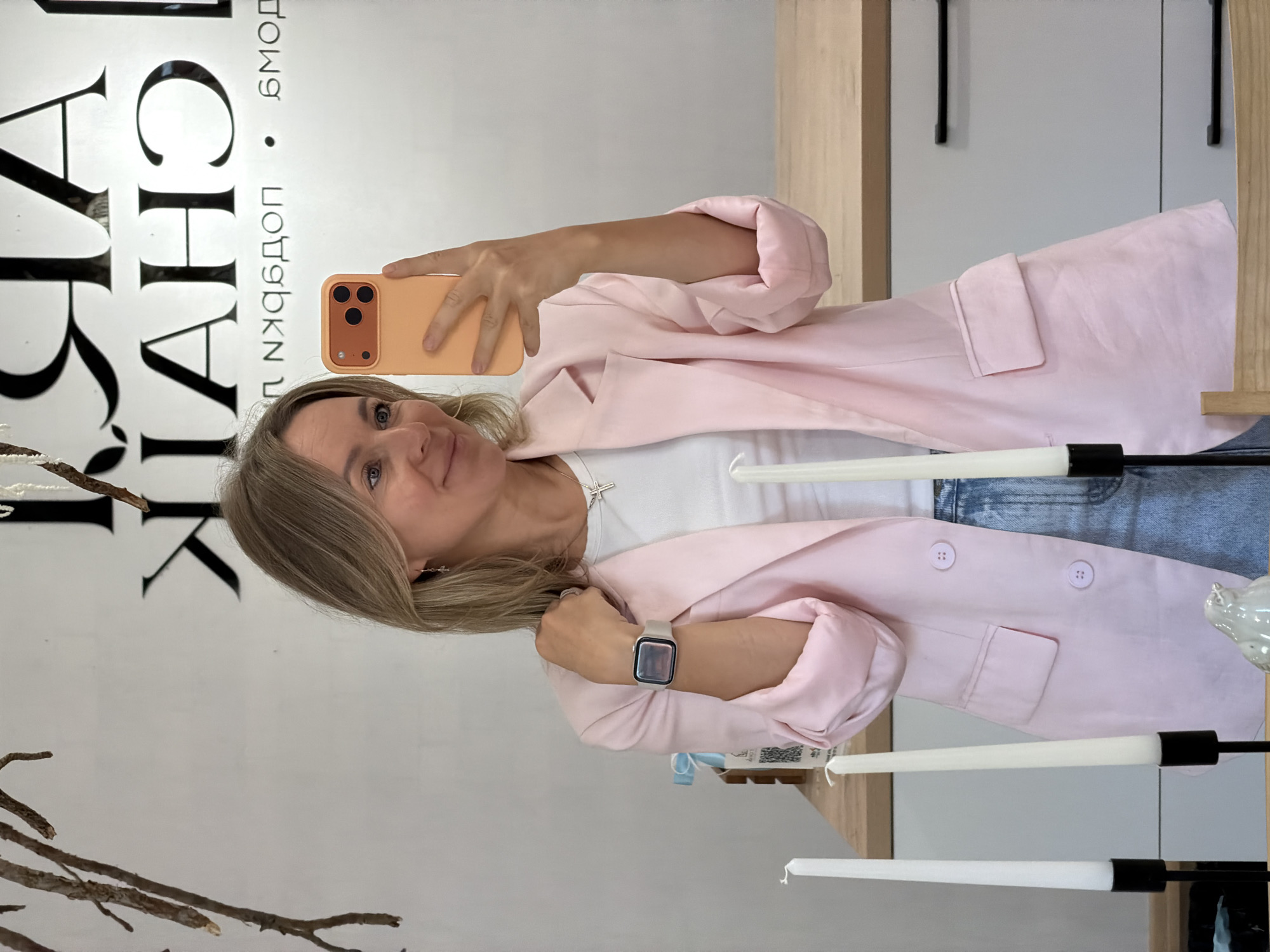 A young blonde woman is taking a selfie in a studio. She is wearing a pink blazer over a white t-shirt and blue jeans. She is holding a peach-colored smartphone in her right hand. In the background, there is a white wall with text that appears to be in Russian. The word "Чайковский" (Chaikovsky) is visible on the wall, indicating the city. There are also two white taper candles in black holders in the foreground, and a small white bird figurine. The scene appears to be set in a modern, minimalist space, possibly a retail store or a photography studio.