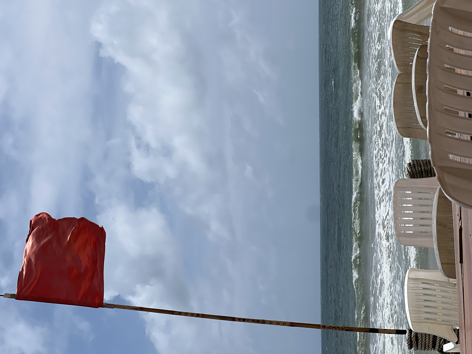 The image depicts a beach scene in Unknown, Unknown. A red warning flag on a pole stands prominently against a cloudy sky. The ocean is visible with waves breaking near the shore. Several white plastic beach chairs are stacked on a wooden deck. The image is taken from a low angle, emphasizing the flag and the sky.