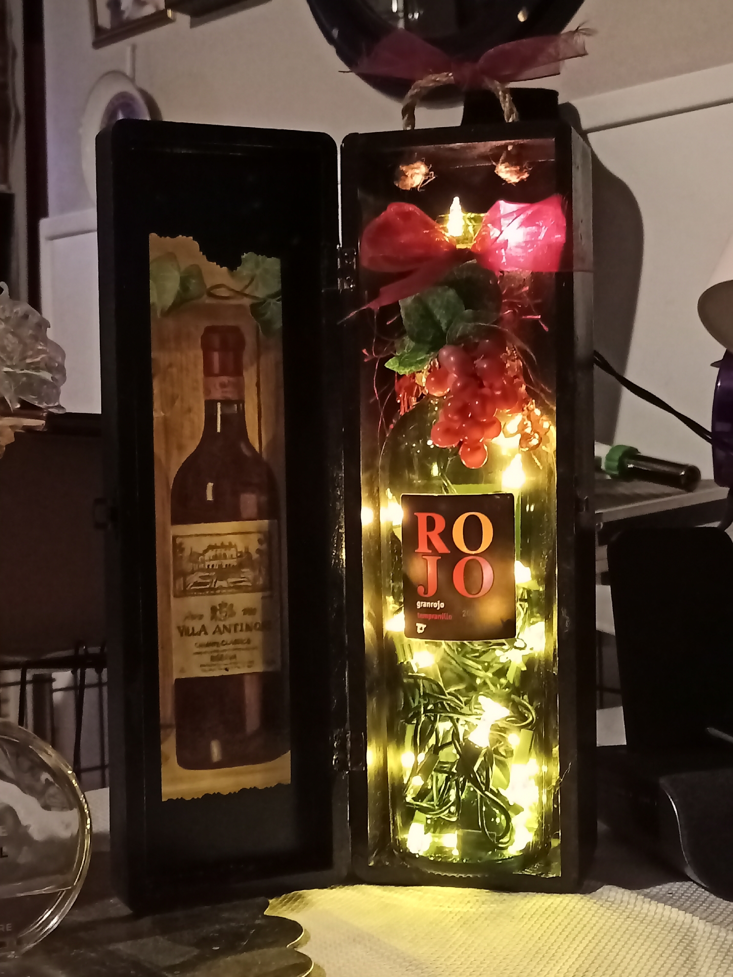 The image shows a decorative wine bottle inside a black wooden box. The box is open, revealing two compartments. The left compartment contains a framed picture of a wine bottle. The right compartment holds a wine bottle decorated with red grapes, a red bow, and illuminated by yellow string lights. The label on the bottle in the right compartment reads "ROJO granrojo tempranillo." The background is blurred, showing what appears to be a room with framed pictures on the wall and various objects on a shelf. There are no people or discernible activity. The location of Amarillo, United States of America is not indicated in the image.