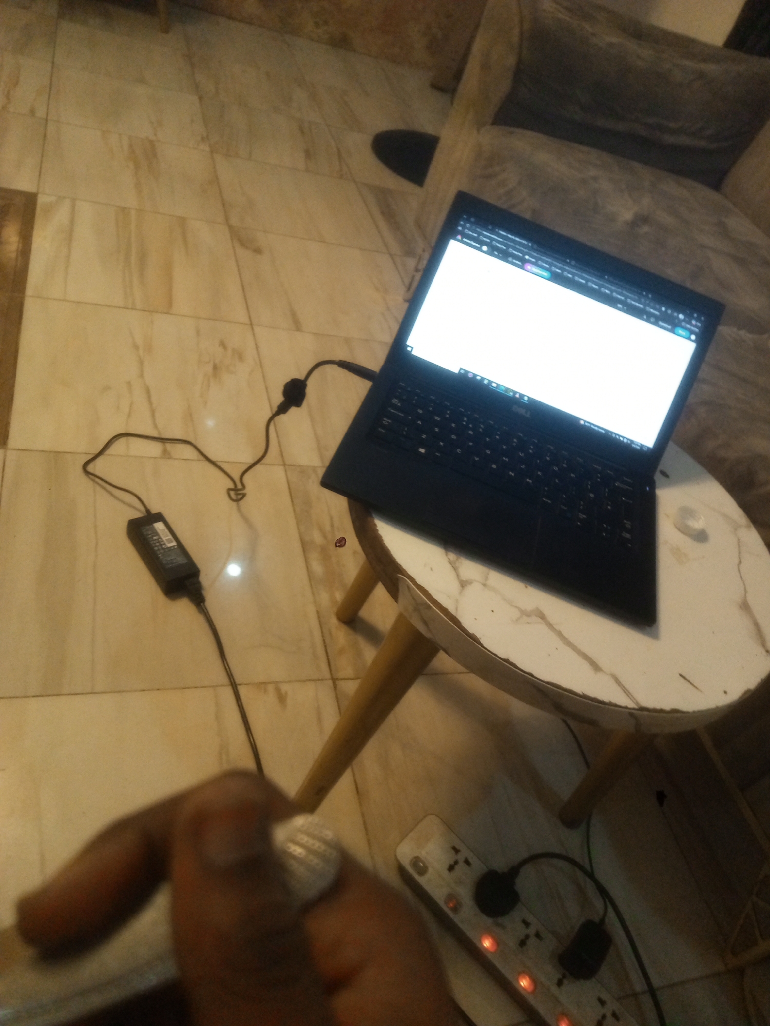 The image shows a laptop, likely a Dell, sitting on a small, round table with a white marble-patterned top. The laptop is open, displaying a screen with what appears to be a web browser or application open. The laptop is connected to a power adapter plugged into an extension cord with multiple sockets and illuminated red power switches. The floor appears to be tiled with light-colored, marble-like tiles. Part of a gray couch is visible in the background. A partially visible hand with a large silver ring on the finger is positioned in the foreground. The setting suggests a domestic environment in Gwarinpa, Nigeria. There is no verifiable way to ascertain the specific location within Gwarinpa from the available information.