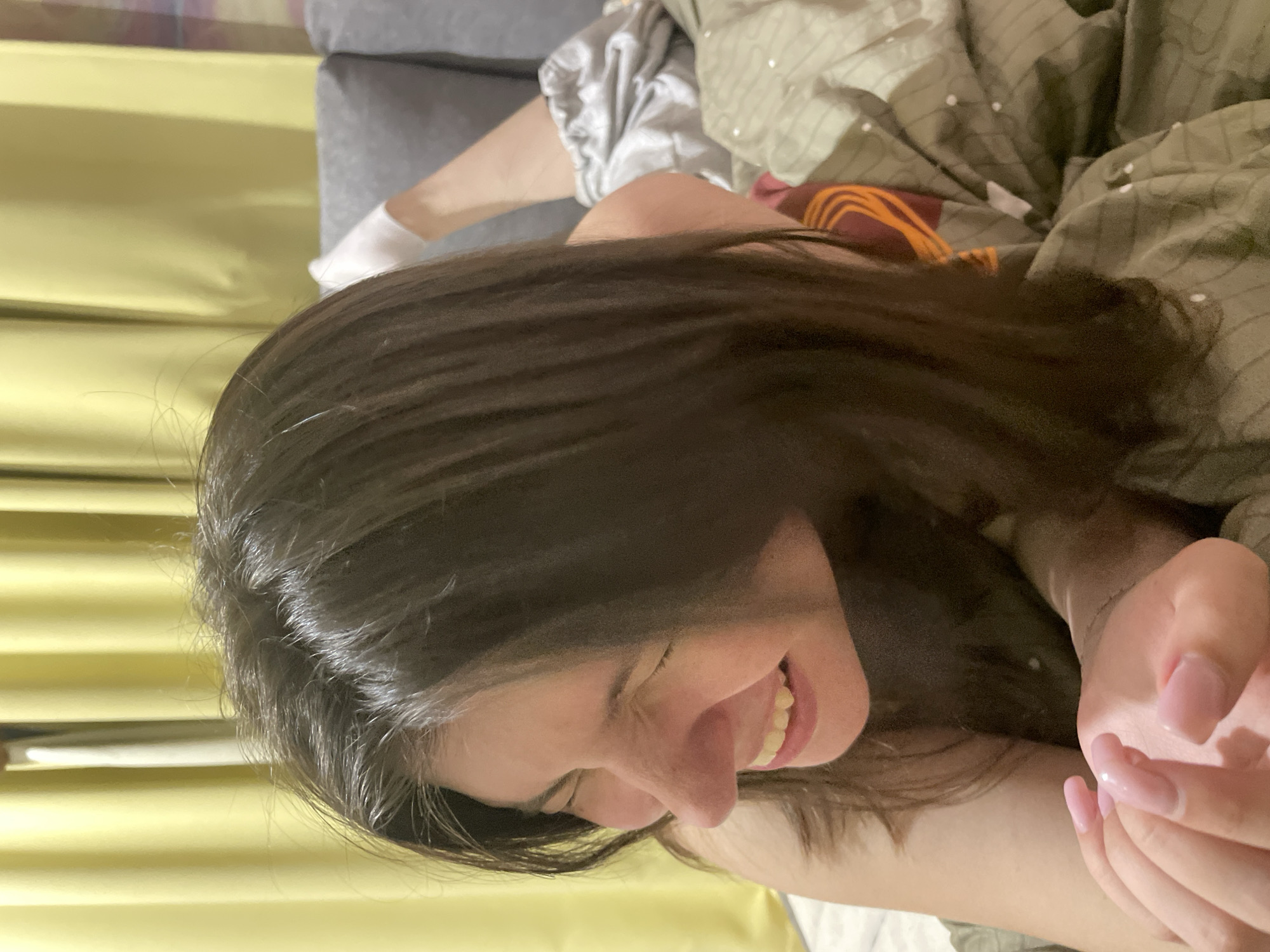 The image captures a close-up of a young woman lying down, with her head tilted and eyes closed in a moment of apparent relaxation or sleep. She has long, dark hair that cascades around her face. Her mouth is slightly open, revealing a hint of a smile, suggesting a sense of peace or contentment. Her skin tone is fair, and her fingernails are painted a light pink.
In the background, the setting appears to be a bedroom or a cozy indoor space. A gray couch is partially visible, along with crumpled bedding in shades of green and brown. The bedding has a subtle pattern of white dots. A hint of yellow fabric, possibly curtains or a wall covering, runs along the left side of the frame. A glimpse of the woman's legs, clad in white socks and shiny, light-colored pajama bottoms, is also visible. The overall ambiance is casual and intimate, likely captured during a quiet moment at home. The lighting suggests it could be daytime or evening, with soft, ambient light. There is no visible text in the image. The location context provided is ЖК «Борисоглебское», Russia, which could indicate the residential area where the photo was taken.