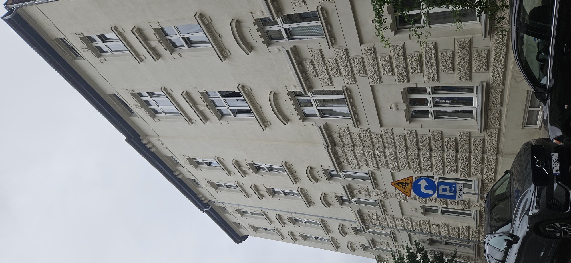 The image shows the facade of a pale yellow, multi-story building in Krakow, Poland. The building has a dark roofline and numerous windows with white frames. Some windows have curtains visible. The facade features decorative stone detailing around the windows and horizontal bands of textured stone.
In the foreground, on the left side, a dark grey Volvo SUV is parked, its license plate reading "WF 362CY". To the right of the SUV, part of another dark car is visible.
There are several traffic signs on poles in front of the building. The topmost sign is a yellow triangle with a black bicycle symbol, indicating a bicycle route. Below that is a blue circular sign with a white arrow pointing right, signifying a mandatory right turn. The lowest sign is a blue square with a white "P" symbol, indicating parking, with the word "Płatny" (meaning "paid" in Polish) and "Koniec" (meaning "end" in Polish) below it, likely indicating the end of a paid parking zone.
The sky is overcast and grey. Some green foliage is visible on the right side of the image, partially obscuring a window on the building's upper floors. The scene appears to be a street in a European city, likely a residential or commercial area, given the building's architecture and the presence of parked cars and traffic signs.