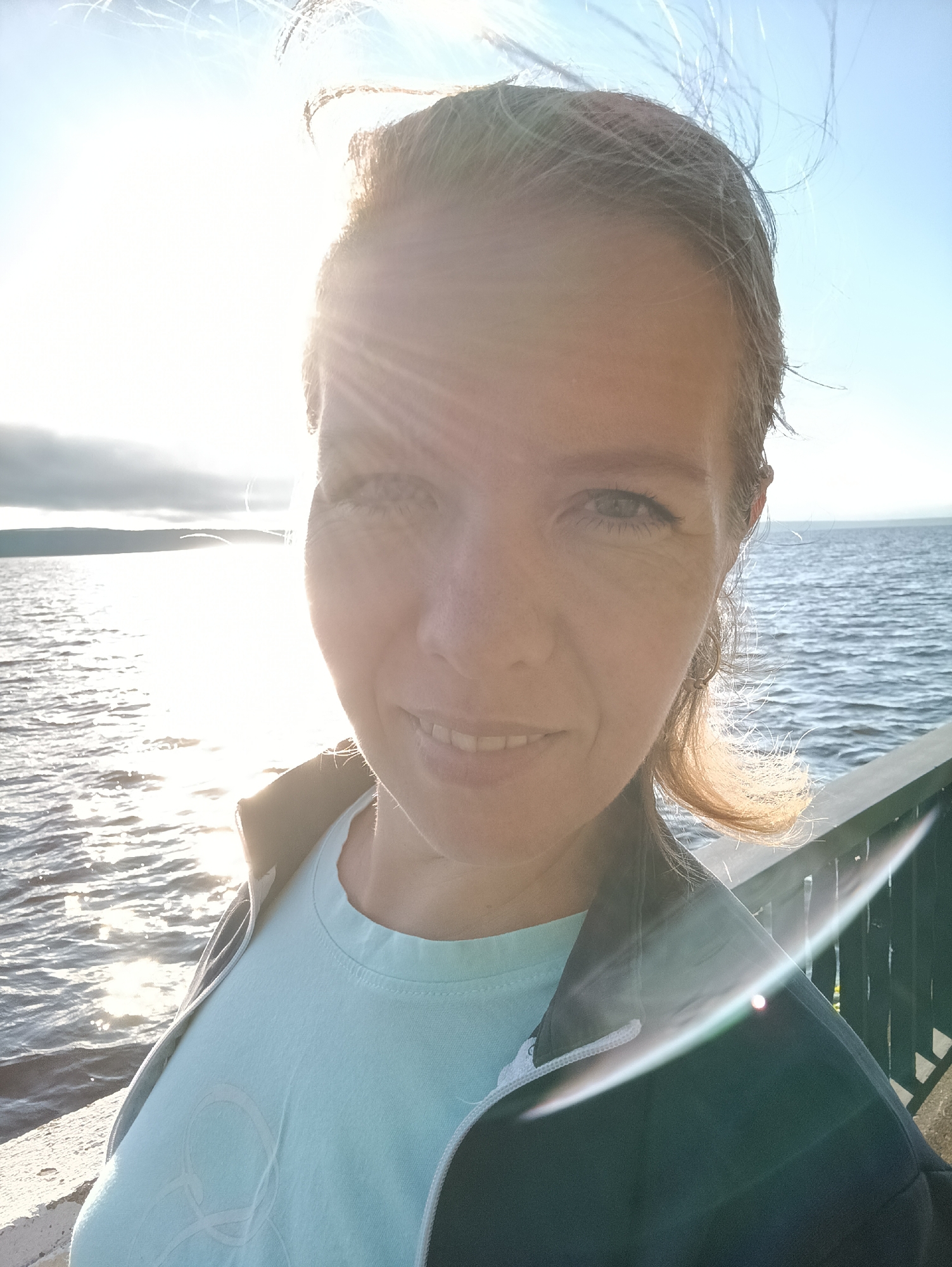 A selfie taken by a woman outdoors. She is facing the camera with a slight smile. The sun is bright and directly behind her, causing lens flare and making the background overexposed in some areas. She is wearing a light blue t-shirt and a dark jacket. Her hair is light brown and windblown. Behind her, there is a body of water, likely the Kama River, and a distant shoreline. The location is on a pier or boardwalk, indicated by a railing visible in the lower right corner. This image was taken in Chaikovsky, Russia.