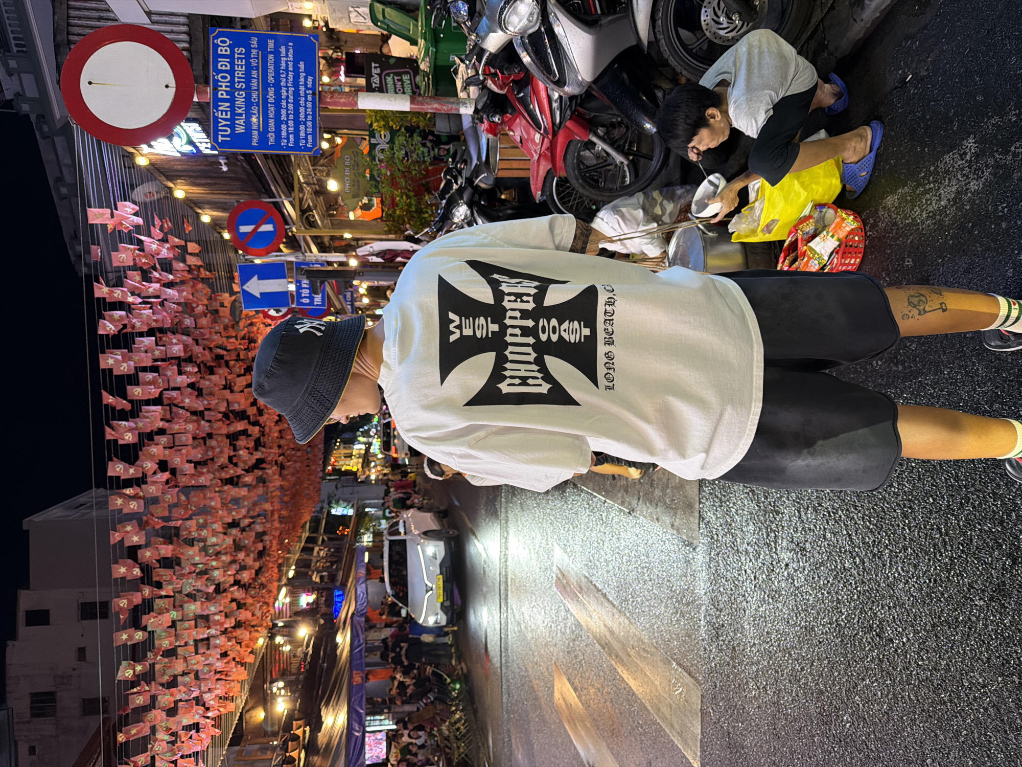 The image depicts a street scene at night in Thuận Hóa Ward, Vietnam. A young man wearing a bucket hat, white t-shirt with a "West Coast Choppers" graphic, and black shorts stands in the foreground, facing away from the camera. His right leg has a tattoo. In the mid-ground, a child in a grey t-shirt and black pants is crouched down, eating from a bowl. Next to the child is a yellow plastic bag and a red plastic basket containing snacks. The street is wet, reflecting the overhead lights and festive red banners hanging across the street. Several motorcycles are parked along the side of the road. Traffic signs, including a "No Entry" sign and a pedestrian crossing sign, are visible. A white car is driving in the background. The overall atmosphere is of a busy, vibrant urban environment at night.