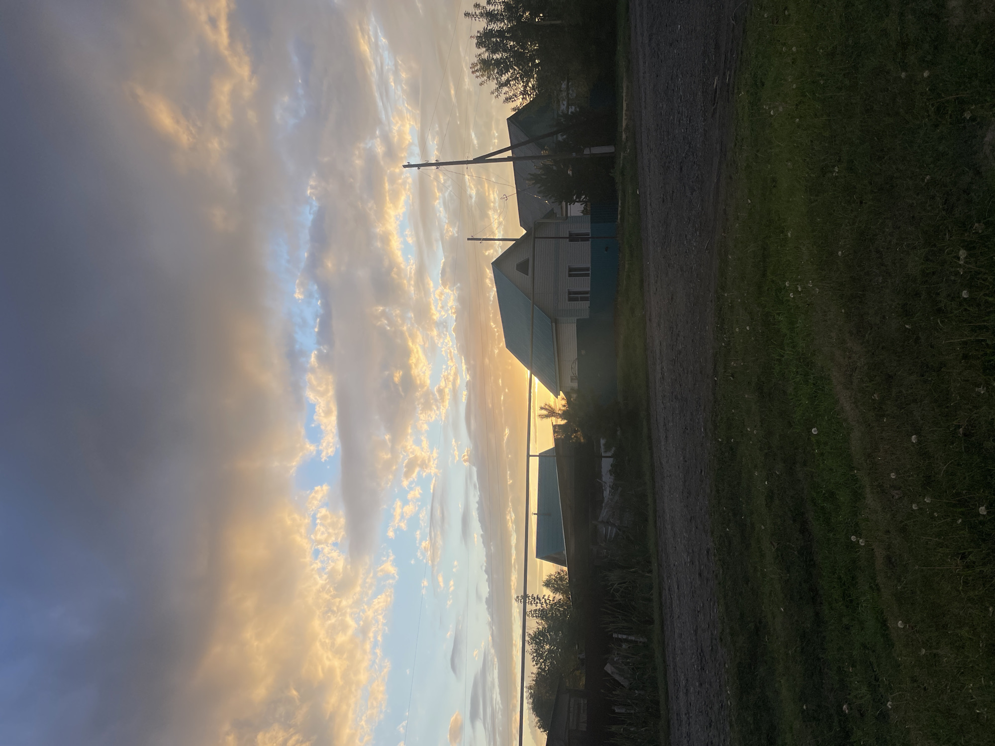 The image shows a rural street scene in the city of Kargat, Russia, during either sunrise or sunset. The sky is filled with dramatic clouds, colored in shades of gray, blue, and warm yellow and orange hues from the sun's low angle. In the background, several houses with teal-colored roofs are visible, suggesting a residential area. A dirt road runs through the foreground, with grassy verges on either side. A few utility poles with overhead wires are also present. The overall atmosphere is tranquil and somewhat overcast, characteristic of a twilight scene in a rural setting.