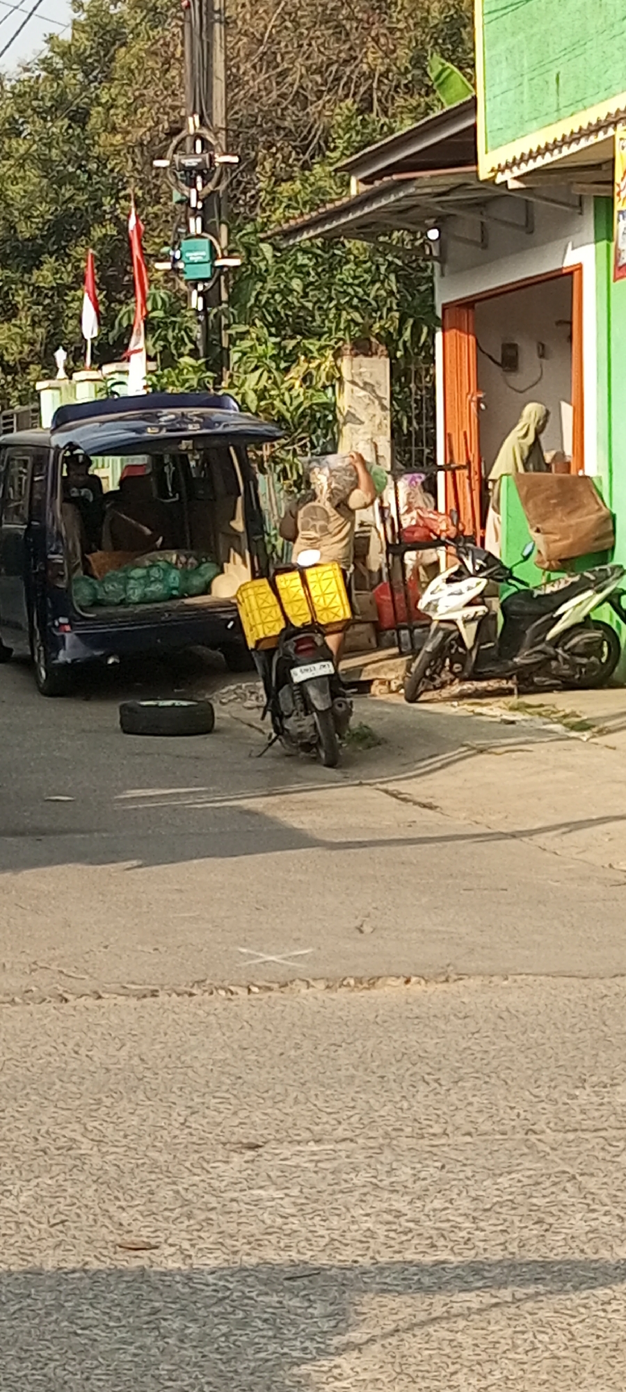 In Setu, Indonesia, a person is seen carrying a large bag of goods over their shoulder, walking away from a dark blue van. The van's tailgate is open, revealing several green plastic bags filled with items. A spare tire lies on the ground next to the van. In front of the van is a motorcycle with two yellow containers attached to the rear. Behind the motorcycle, a white scooter is parked. The scene unfolds on a paved road in front of a building with a green facade and an orange-framed doorway. Overhead, trees with sparse foliage suggest a dry season, and a utility pole with traffic lights and two Indonesian flags is visible. The ground is cracked and dry, with faint white markings.