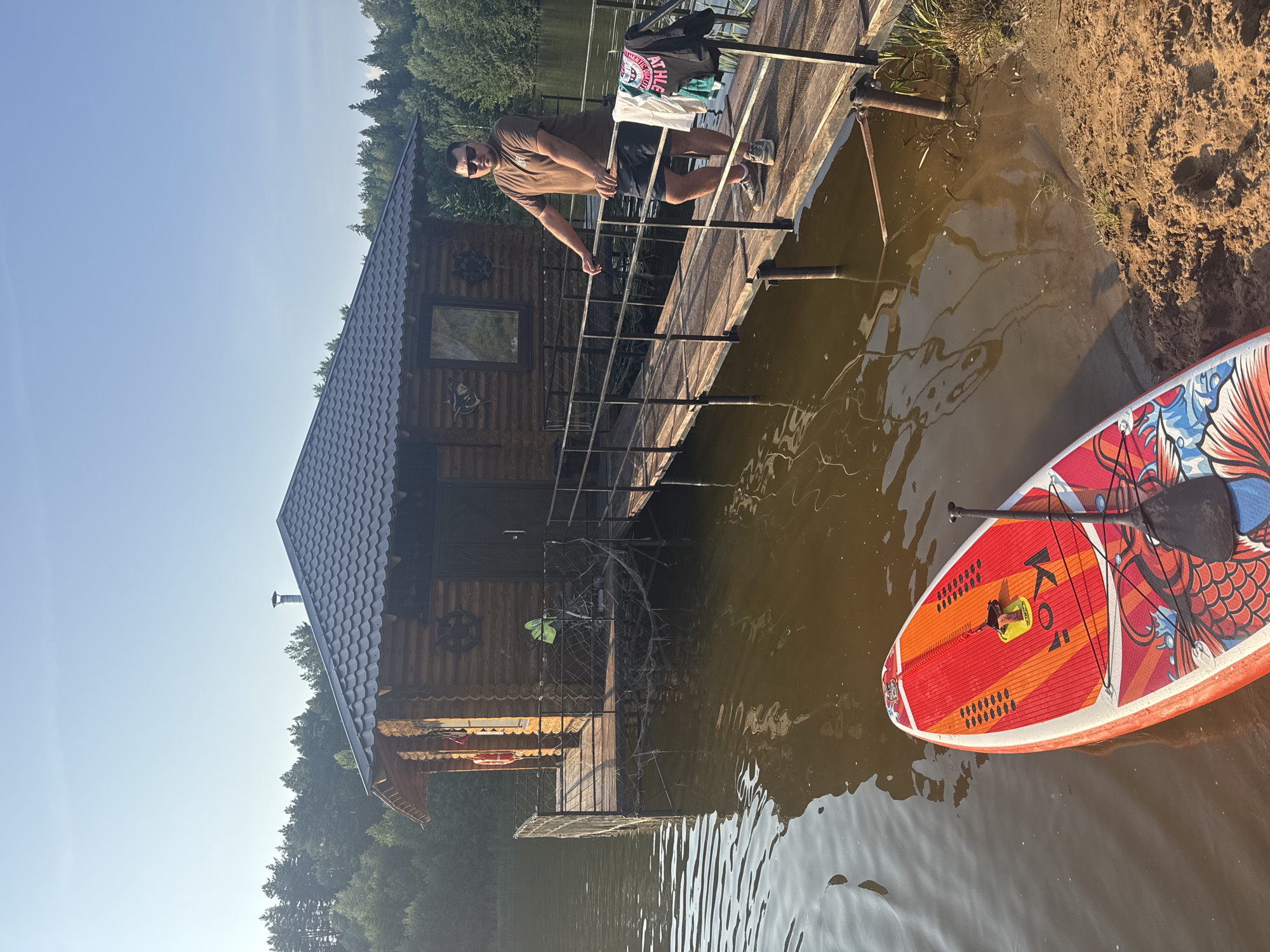 A man stands on a wooden pier leading to a log cabin on a body of water. The cabin has a dark, shingled roof and a wooden door. There is a paddleboard with a red and orange design floating in the water in the foreground. The man is wearing a brown t-shirt, black shorts, and sneakers. He is looking towards the camera. There are trees in the background. The scene appears to be in Prikamskii, Russia.