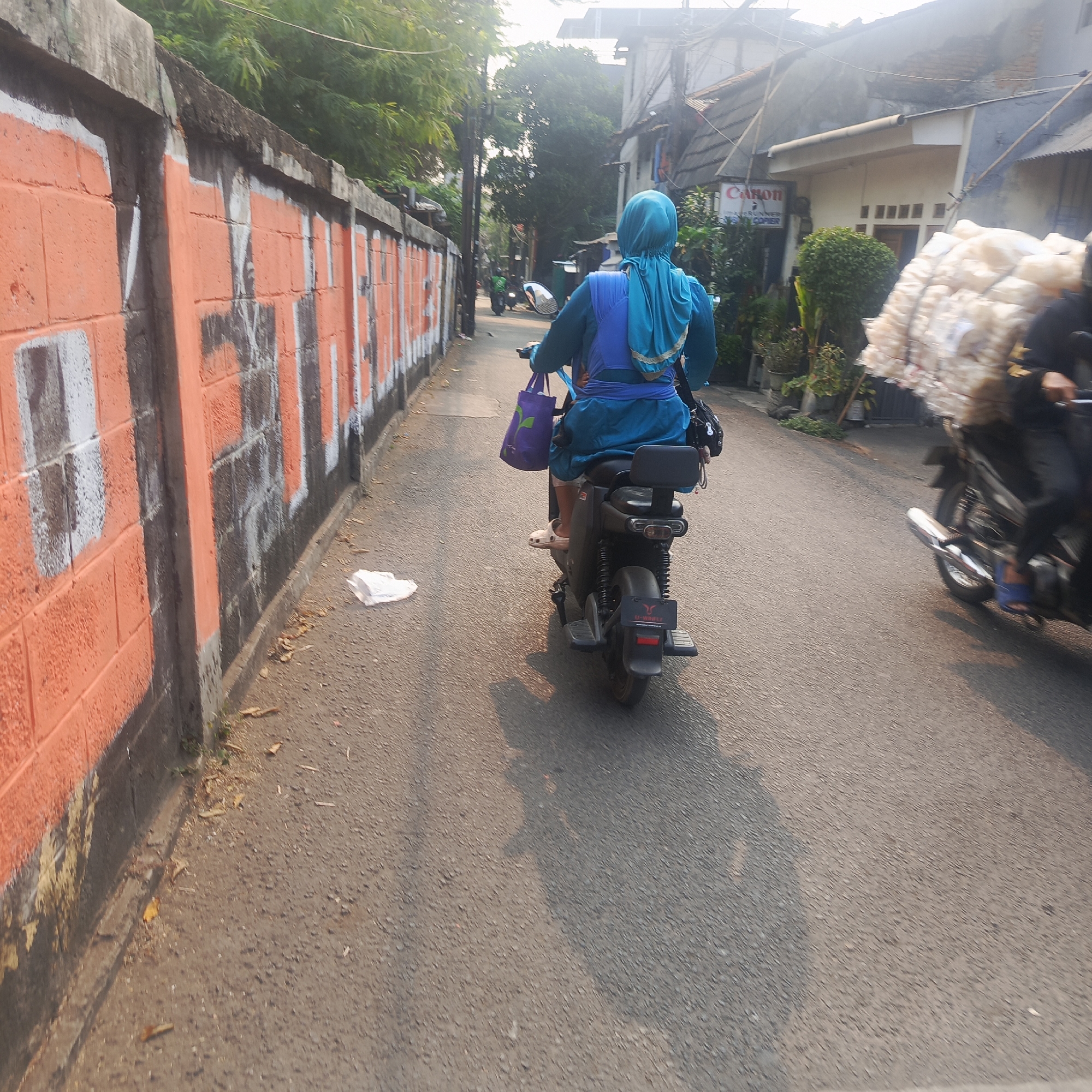 The image depicts a street scene in South Jakarta, Indonesia. In the foreground, a person wearing a blue hijab and blue clothing is riding an electric scooter. They are carrying a purple bag and appear to have a child in a carrier strapped to their back. The scooter has a black seat and body with a red taillight and a visible logo. To the left of the street is a textured orange brick wall with black and white graffiti. The road is paved and appears to be an alleyway or narrow street with buildings and trees lining the sides. In the middle ground, another person on a motorcycle is visible, carrying a large stack of white material, possibly food items or packaging. A sign for "Canon" is visible on a building in the background. The lighting suggests it is daytime with sunlight casting shadows.