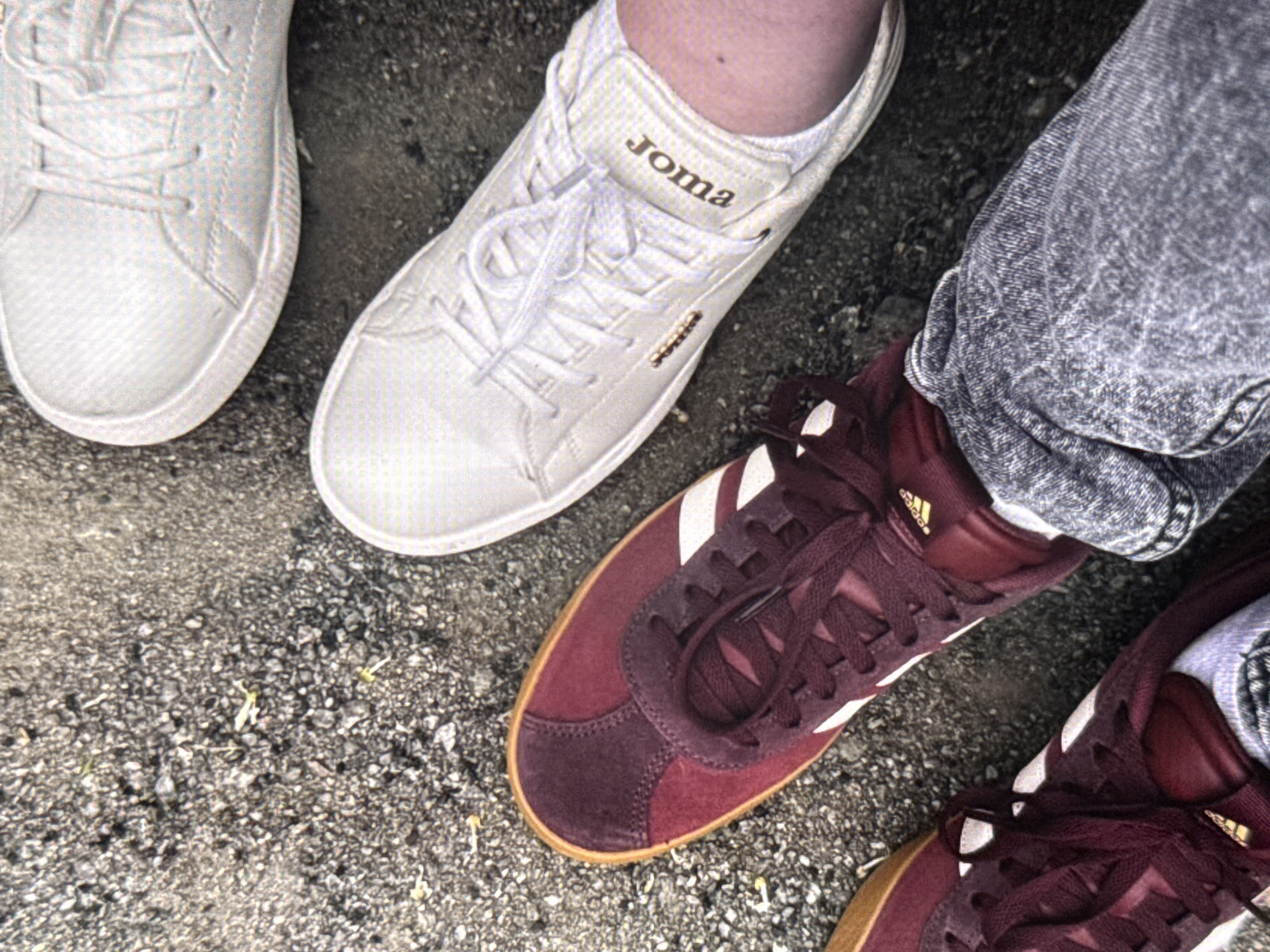 The image depicts an overhead view of four feet belonging to two individuals standing on a rough, dark grey ground surface, consistent with asphalt or fine gravel. The specific location within the city of Kyiv, Ukraine, cannot be determined from the visual information provided.
The individual on the left is wearing a pair of dark-wash or acid-wash denim jeans and a pair of dark burgundy/maroon low-top sneakers featuring white stripes on the side and a light brown gum sole. A small golden Adidas logo is visible on the inner side of one of these shoes. The laces are tied in a standard cross pattern.
The individual on the right is wearing two white low-top sneakers. The shoe closer to the foreground displays the brand name "Joma" in golden lettering on its tongue, oriented upside down from the viewer's perspective. A small golden metallic detail is also present near the laces of this shoe. The laces are white and tied.
The ground surface is composed of small, dark grey stones and sand-like particles. Scattered across the surface are a few very small, light green plant sprouts or debris.