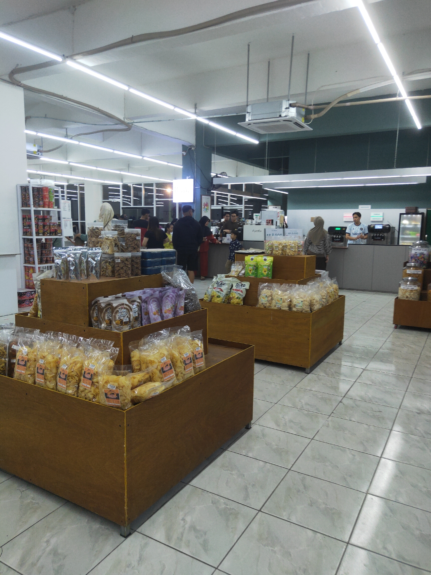 The image depicts the interior of a small grocery store or minimarket in Sintang, Indonesia. The store features a tiled floor and is illuminated by fluorescent lights in the ceiling. Several wooden display stands are filled with bagged snacks and food items, some of which are clearly visible in plastic packaging. In the background, a counter with a cash register and possibly a coffee machine is present. Two employees are visible behind the counter, and several customers are browsing the aisles or waiting at the counter. Shelves stocked with various products are visible on the left side of the image. The overall scene suggests a typical retail environment.