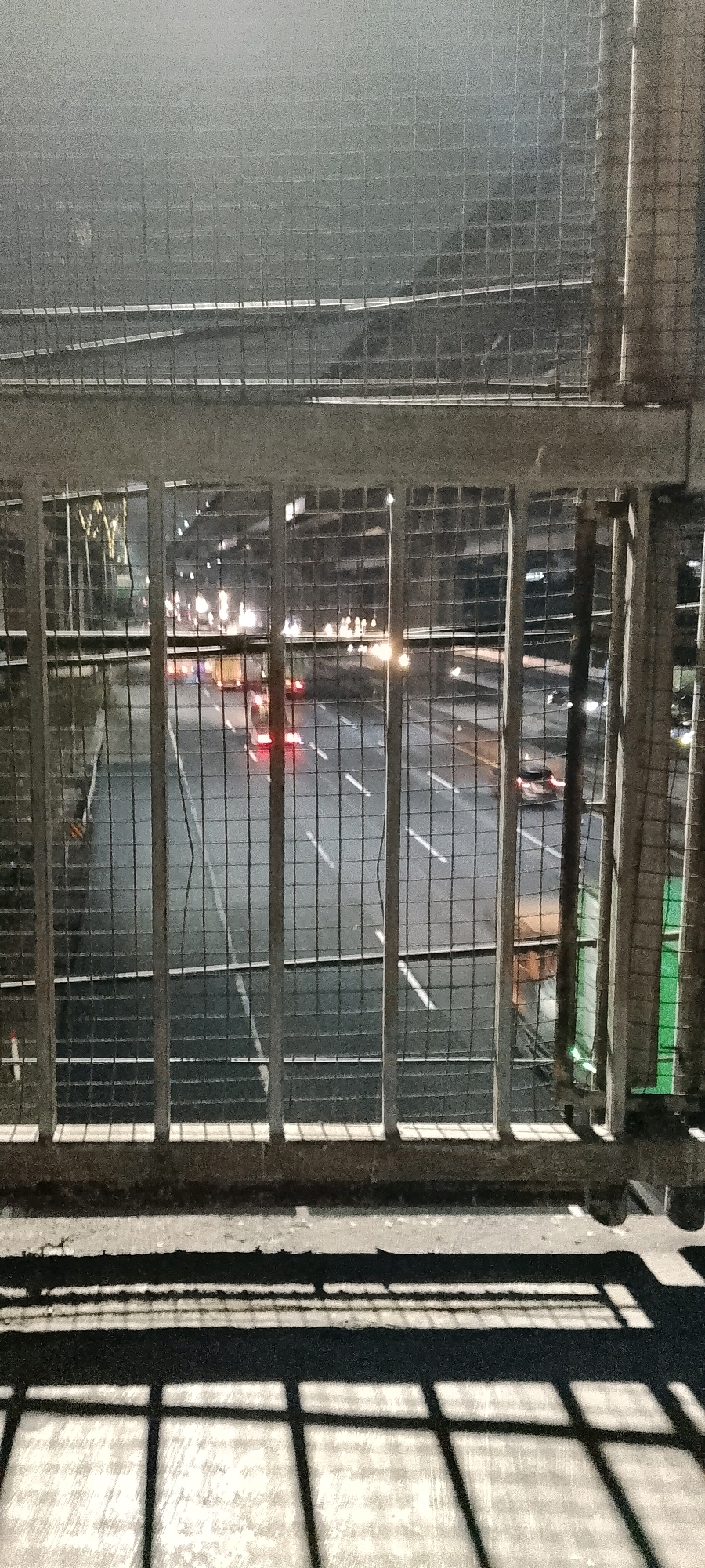 An overpass view of a highway at night in Bekasi, Indonesia. A chain-link fence runs across the foreground, separating the viewer from the scene below. The highway is wet and reflects the lights of passing vehicles, which appear as streaks of red, white, and yellow. Buildings line the sides of the highway, illuminated by streetlights and interior lights. The overall atmosphere is dark and somewhat hazy. The concrete overpass structure itself is visible in the foreground, with shadows cast from the fence onto the walkway.