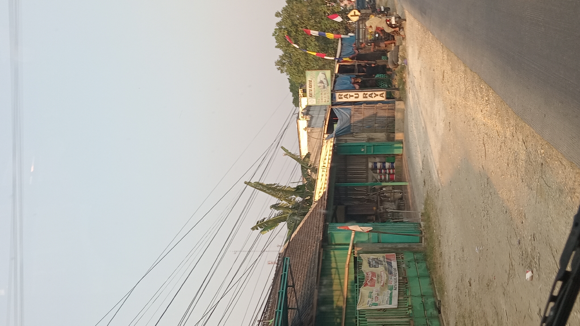 The image shows a street view in Kunduran, Indonesia, with a row of small businesses lining the left side of the road. The businesses appear to be an auto repair shop and possibly a store selling paint and auto parts. A banner on the left advertises "AKU RAYA MOTOR" with images of car parts. Another sign reads "RATU RAYA". There are several motorcycles parked in front of the establishments. In the foreground, a portion of a vehicle's dashboard is visible, suggesting the photo was taken from inside a car. The sky is clear and bright, indicating daytime. The road is paved with asphalt.