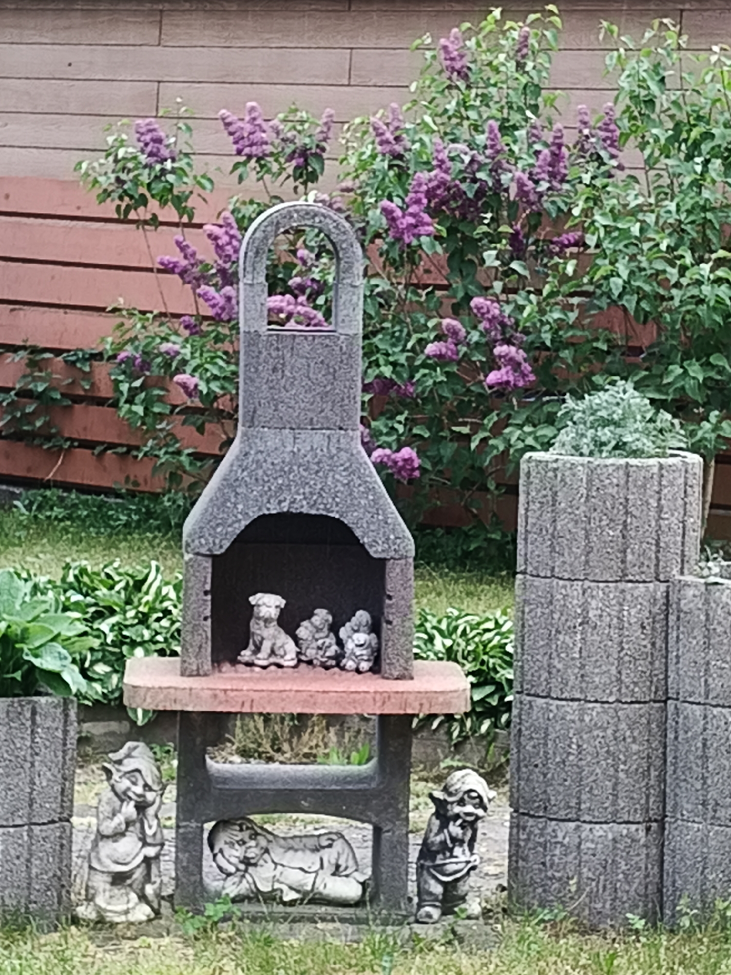 This image depicts a garden scene in Unknown, Unknown, featuring various decorative elements. In the center, a gray, granular stone or concrete garden structure resembles a miniature fireplace or shrine. The upper section of this structure has an arched opening containing three light-colored figurines: one resembling a small dog or bulldog, and two smaller, less distinct figures. Below this opening, a flat shelf separates the upper and lower sections. The lower section contains a single, light-colored figurine of a reclining, large-headed creature, possibly a gargoyle or fantastical animal.
To the left of the central structure, a light-colored, gnome-like garden statue with a pointed hood is positioned, appearing to hold its hand to its chin. Another similar gnome-like figure stands to the right of the central structure's base. Further to the right, a tiered structure of stacked, rough-hewn concrete blocks serves as a planter, with a small, light-green, bushy plant growing in the top section. A partial view of a similar concrete block structure is visible in the lower-left foreground.
The ground is covered with green grass and various broad-leafed green plants, including what appears to be hostas, particularly on the left side of the scene. In the background, a horizontal wooden fence, stained a reddish-brown color, runs across the frame. Numerous green bushes with clusters of purple flowers, consistent with lilacs, are visible both in front of and behind this fence, dominating the upper and mid-background. The scene is illuminated by natural daylight. No human figures are present.