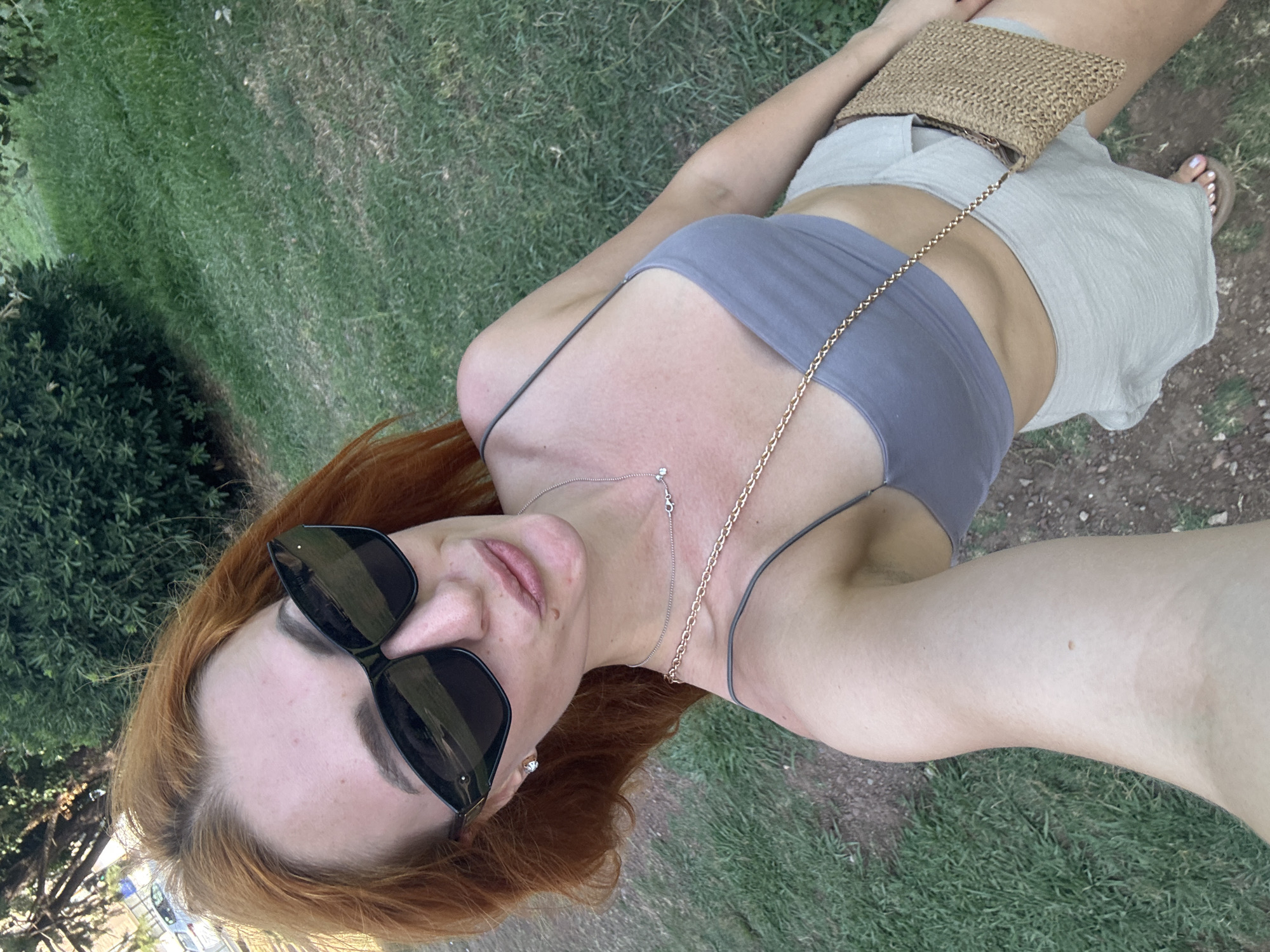 A woman with red hair and sunglasses is standing outside, possibly in a park or garden, with grass and trees in the background. She is wearing a grey crop top and light-colored shorts. She is holding a straw-woven purse. She has a necklace on and is wearing earrings. The woman is looking at the camera and appears to be taking a selfie. The image appears to be taken during the daytime. The location is identified as Budva, Montenegro.