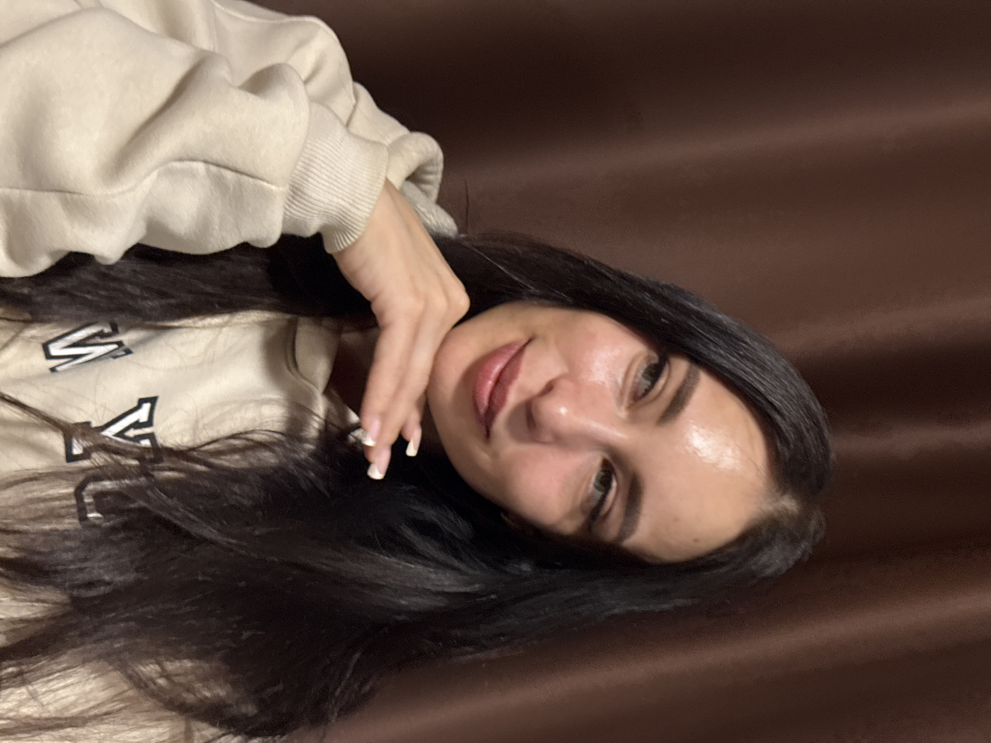 The image captures a young woman with long, dark hair, lying down and looking towards the camera with a gentle smile. Her head is tilted, and her right hand, adorned with neatly manicured nails (light-colored with white tips), rests near her chin. She is wearing a light-colored, possibly beige or off-white, hooded sweatshirt with ribbed cuffs. A dark letter, resembling a "V," is partially visible on the front of her hoodie, along with other faint, illegible markings.
The woman has light-colored eyes, well-defined eyebrows, and wears light lipstick. Her expression appears relaxed and pleasant. The background is a solid, dark brown or maroon color, likely a curtain, wall, or piece of furniture, providing a soft, uniform backdrop. The lighting suggests an indoor setting. There are no visible cues for the time of day or specific weather conditions. The overall mood is calm and casual. The image context points to Chaikovsky, Russia, as the location.
