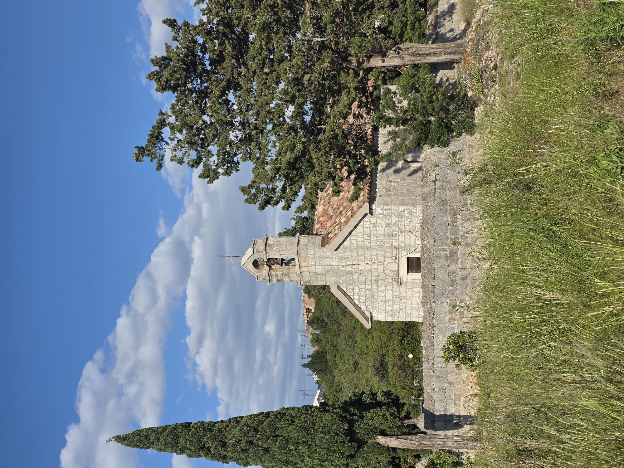 This image presents a serene outdoor scene in Luštica, Montenegro, centered around a traditional stone church situated on a hillside. The church itself is constructed from light-colored, possibly grey or tan, stone blocks and features a distinctive reddish-brown tiled roof. A prominent bell tower, or campanile, rises from the front of the church, topped with a small cross and containing an arched opening. A dark wooden door and a small window with dark shutters are visible on the side of the building.
The church is surrounded by lush green vegetation. A large, densely foliated green tree, likely a pine or cypress, partially overhangs the church from the left, its branches extending into the sky. Another tall, slender cypress tree stands in the lower left portion of the frame. The foreground and the right side of the image are covered with an abundance of wild, tall green grass and scattered shrubs, giving the scene a natural, somewhat untamed feel. A thick tree trunk is also visible lying horizontally near the grassy area on the right.
The sky above is a vibrant blue, adorned with scattered, fluffy white cumulus clouds, indicating bright and clear weather. In the distance, beyond the church, rolling hills covered in dense green forest are discernible, with some faint man-made structures or power lines visible on the horizon. The scene is bathed in bright daylight, suggesting either morning or early afternoon. There are no people present in the image, nor is any visible text. The overall impression is one of peacefulness, historical charm, and natural beauty.
