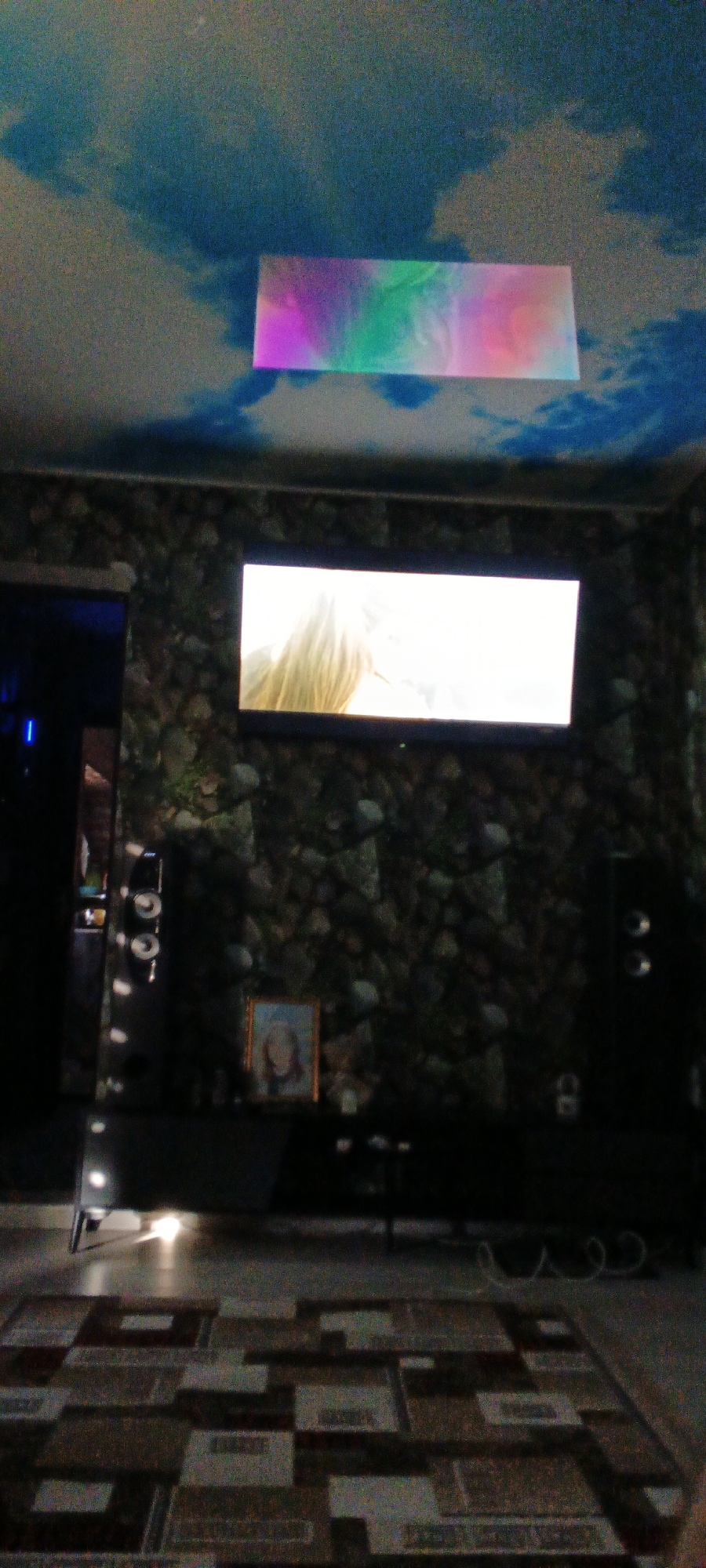 The image depicts an interior scene, likely a living room, with a television mounted on a stone-patterned wall. Above the television, a rectangular projection of colorful light, possibly from a projector, is visible on the ceiling which is painted with a sky and cloud motif. The room contains a media console below the television, on which a framed picture is displayed. Two speakers are visible, one on the left and one on the right side of the television. The floor is covered with a patterned rug. The overall lighting is dim, suggesting the image was taken in the evening or in a darkened room. There are no people visible in the image. The location is specified as Mustakillik, Uzbekistan.