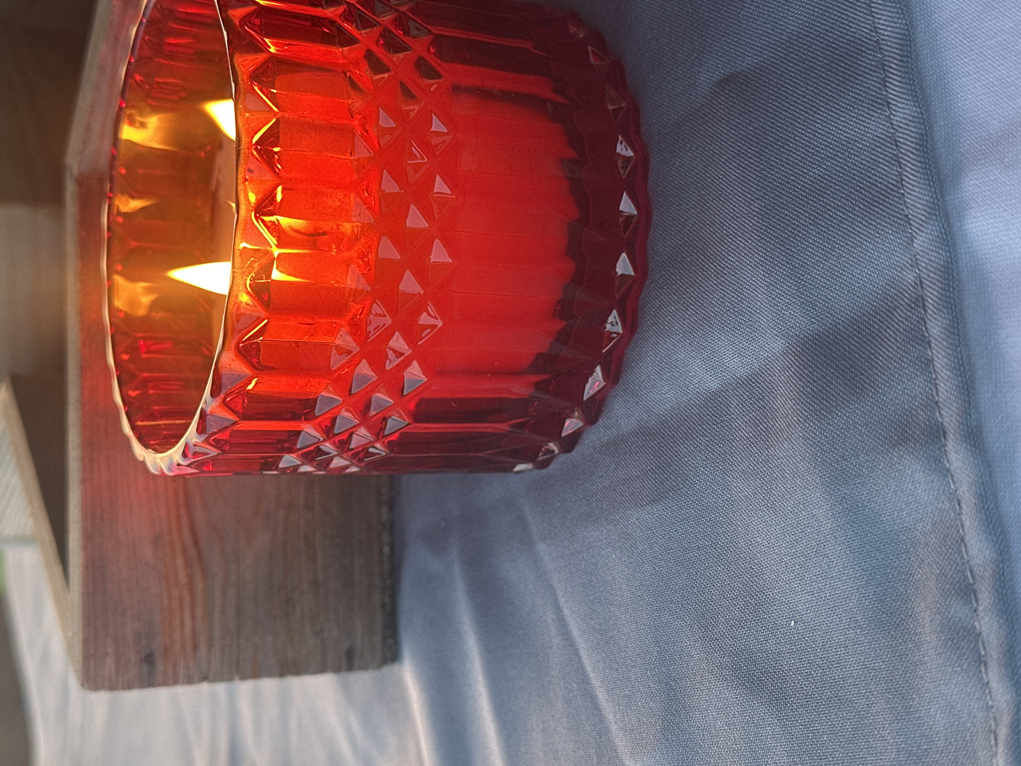 A close-up, eye-level shot focuses on a red, faceted glass jar holding a lit candle with two visible flames. The candle is situated on a textured, muted blue cloth. To the left, a portion of a rough-hewn wooden object, possibly a crate or box, is visible, also resting on the blue cloth. The lighting is warm and soft, suggesting natural light, possibly from a sunset or sunrise. The image does not contain any information about the city of Kharnavy, Russia, or any people or actions.