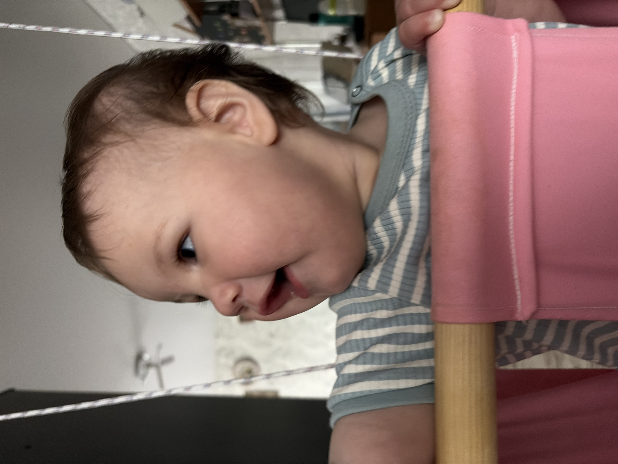 A baby in a blue and white striped shirt is partially visible, holding onto what appears to be a pink object or a piece of furniture with a wooden support. The background is blurry but seems to be an interior space. There is no indication of the location being Novopokrovskaya, Russia.