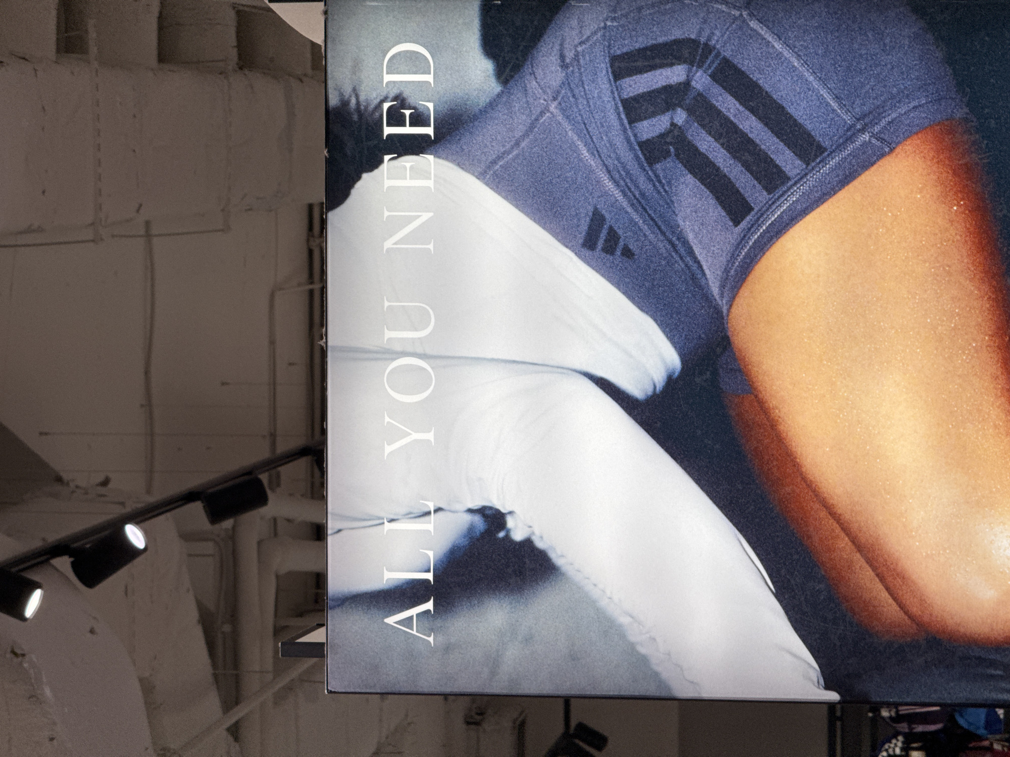 This media file captures a large, vertically oriented advertising banner prominently displayed indoors in Sofia, Bulgaria. The banner features a close-up, dynamic shot of a person, likely an athlete, from the mid-torso down. The individual's skin, visible on their thigh and hip area, appears to be glistening with sweat, suggesting recent physical exertion.
The person is wearing dark blue Adidas athletic shorts or compression wear with visible black triple stripes on the side, a signature Adidas design. A smaller Adidas logo (three bars) is also discernible on the apparel. Below the shorts, white athletic leggings or compression wear cover the lower leg, visible partially.
The text "ALL YOU NEED" is written vertically in white capital letters along the left side of the banner, positioned alongside the person's body.
The background of the image, visible to the left and above the banner, shows parts of an interior space. This area features light-colored, possibly concrete or painted brick, ceiling and wall structures. Exposed pipes and a track lighting system with multiple white lights are visible, indicating an industrial or modern commercial setting, such as a gym, sports store, or retail space. The lighting within the space is artificial, casting a subdued, even tone on the background elements. The overall impression is one of fitness, strength, and athletic pursuit, emphasized by the advertising.