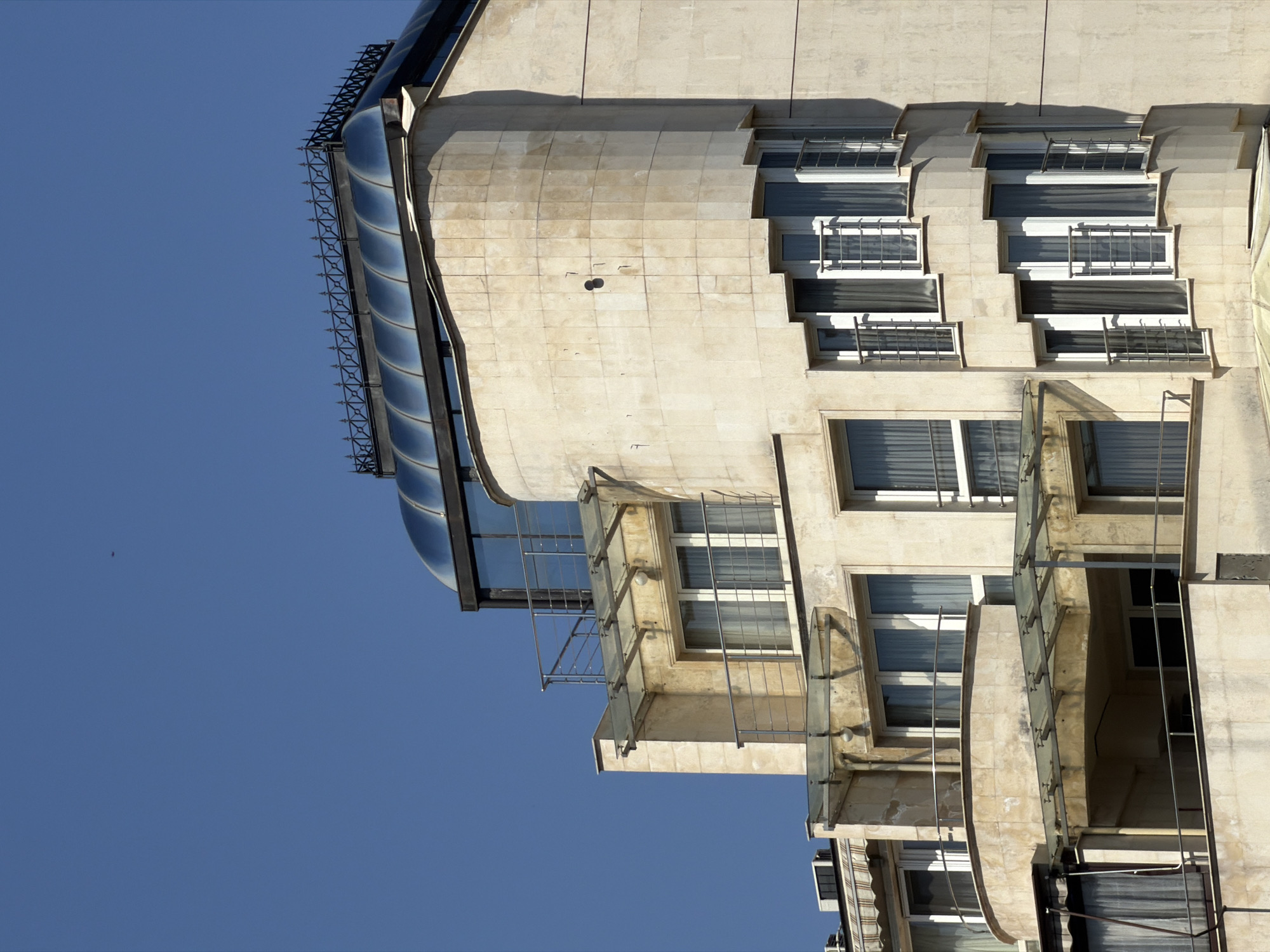 A partial view of a building in Sofia, Bulgaria is captured from a low angle against a clear, blue sky. The building features a mix of architectural styles with curved glass elements on the upper floors, contrasting with the more conventional, box-shaped windows and balconies lower down. The facade appears to be constructed from light-colored stone or concrete. Some balconies have glass panels and metal railings, while others seem to be recessed or enclosed. The lighting suggests a sunny day.