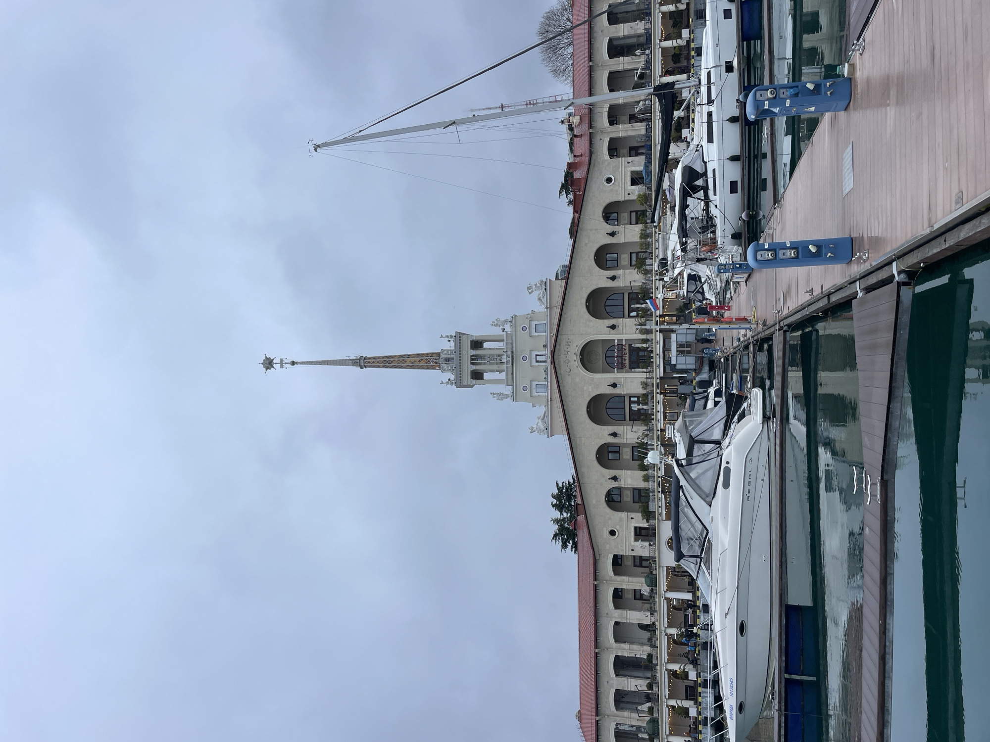 The image displays the Sochi Seaport building and its adjacent marina in Sochi, Russia, under an overcast sky. In the foreground, a dark brown wooden floating dock extends across the lower portion of the frame, with two blue utility pedestals equipped with power and water connections visible on the right side. The water in the marina is a calm, deep teal color, reflecting the boats and dock.
Multiple vessels are moored in the marina. On the left, a white motor yacht, identified as "REGAL 2" with registration "ОФИЦИАДА 10128585," is docked. It features a black canvas bimini top. To the right, a white sailboat with a tall mast, partially visible, is also moored. Fenders are positioned along the sides of the boats.
The central background is dominated by the grand, multi-story Sochi Seaport building. This structure features a light cream-colored facade, a red-tiled roof, and classical architectural elements including arched windows and doorways on its lower levels, and sculptures on its upper sections. The letters "СОЧИ" are visible on the building's facade. A prominent, ornate central spire, topped with a star-like finial, rises from the building. A Russian flag is visible on a flagpole near the building's base. Small groups of individuals are discernible on the docks and along the promenade in front of the building. Coniferous trees are visible to the left behind the building, and a bare deciduous tree is visible to the far right. A distant, red-tipped tower is partially visible on the right horizon, behind the sailboat's mast.