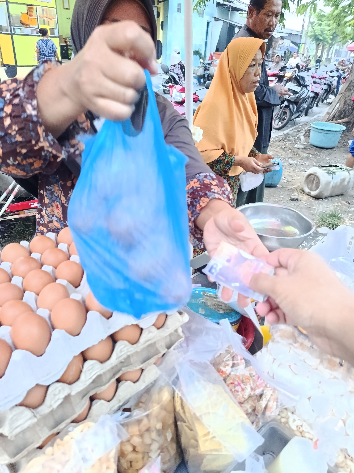 A medium shot captures an outdoor marketplace scene in Keboansikep, Indonesia, with a focus on a transaction. In the foreground, a person wearing a patterned long-sleeved shirt and a dark hijab is handing a blue plastic bag, seemingly filled with items, to another individual whose hands are receiving it, holding a small stack of colorful banknotes. Directly to the left of the blue bag, a tray filled with brown eggs is visible. Behind the primary subjects, another woman wearing an orange hijab stands, looking to the side, while a man with a mustache and dark clothing stands behind her. In the background, various motorcycles are parked, and a blue plastic tub is on the ground. The overall environment suggests a bustling street market with stalls and vendors.