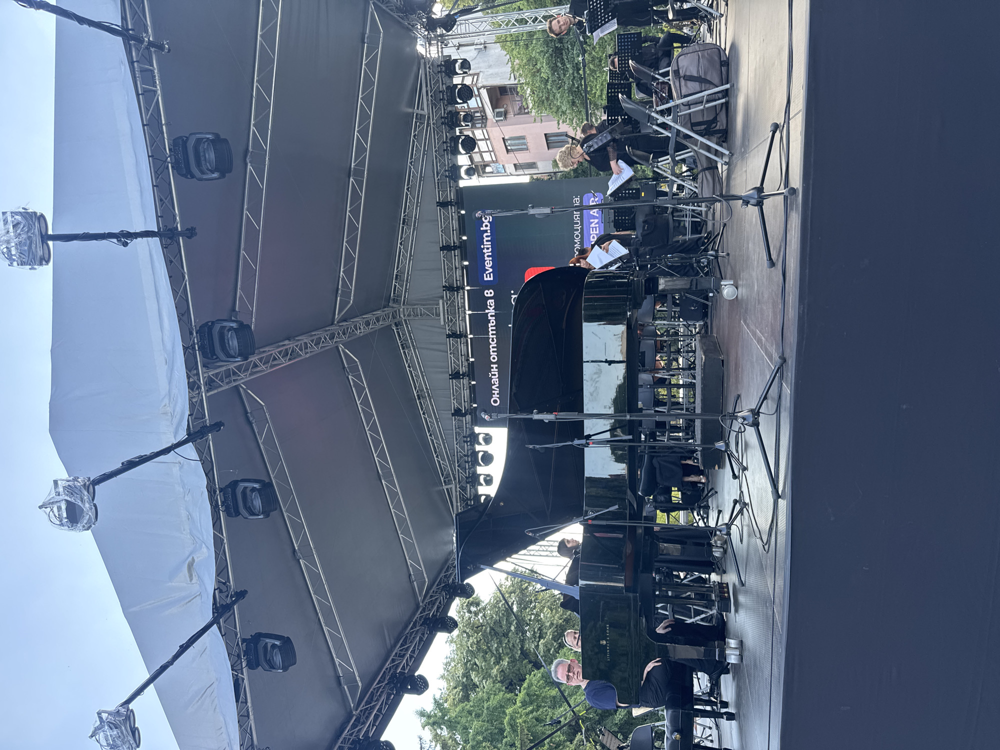 The image depicts an outdoor concert stage in Plovdiv, Bulgaria. On the stage, a grand piano is prominently featured, with several musicians gathered around it. Some individuals are seated at the piano, while others stand, holding sheet music. A large screen is visible behind the performers, displaying text in Bulgarian and the website "Eventim.bg," indicating an organized event. Stage lighting and sound equipment are set up overhead. The background reveals trees and buildings, suggesting an urban setting. The overall scene captures a moment of musical performance.