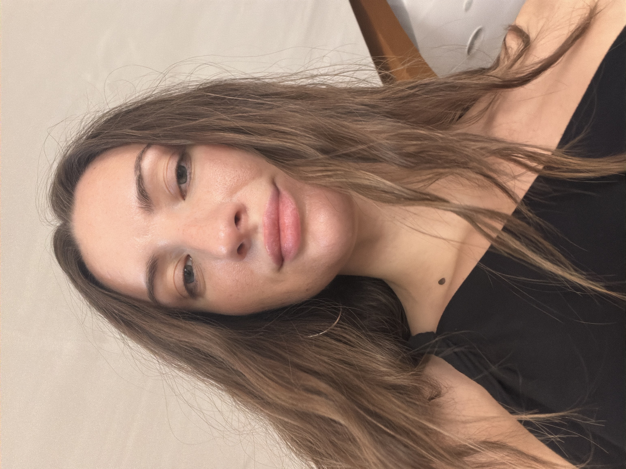 A close-up, low-angle shot captures a woman from the chest up, indoors, against a pale, textured wall. She has long, brown, wavy hair that falls over her shoulders and across her face. Her skin appears to have a dewy finish, and she is wearing a black, one-shouldered top. A small, dark mole is visible on her upper chest, just below her collarbone. She is also wearing a thin, silver hoop earring in her left ear. The background is out of focus, but suggests an indoor setting with a hint of wooden furniture on the right side. The lighting is soft and diffused, possibly from an artificial source. The image does not contain any identifiable landmarks or elements that would indicate a specific location within Dahab, Egypt.