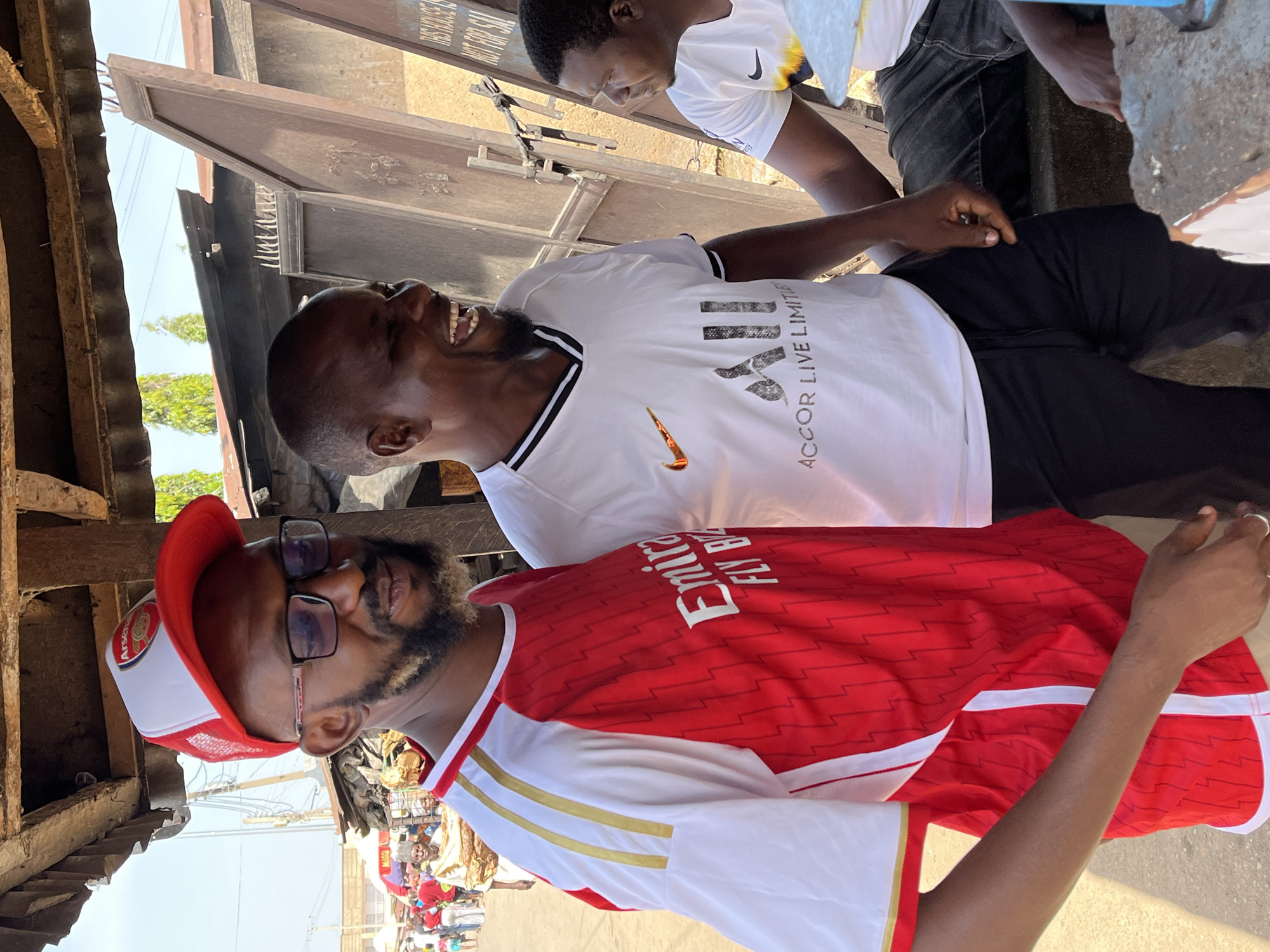 Three men are captured in an outdoor setting. The man in the foreground wears a red and white Arsenal jersey and a red and white cap, while the man behind him wears a white t-shirt. The third man is partially visible behind the second. The location appears to be a bustling street in Jos, Nigeria, as evidenced by the presence of other people and structures in the background. The men are interacting, with at least one of them smiling or laughing. The overall setting suggests an informal gathering or social interaction in an urban environment.