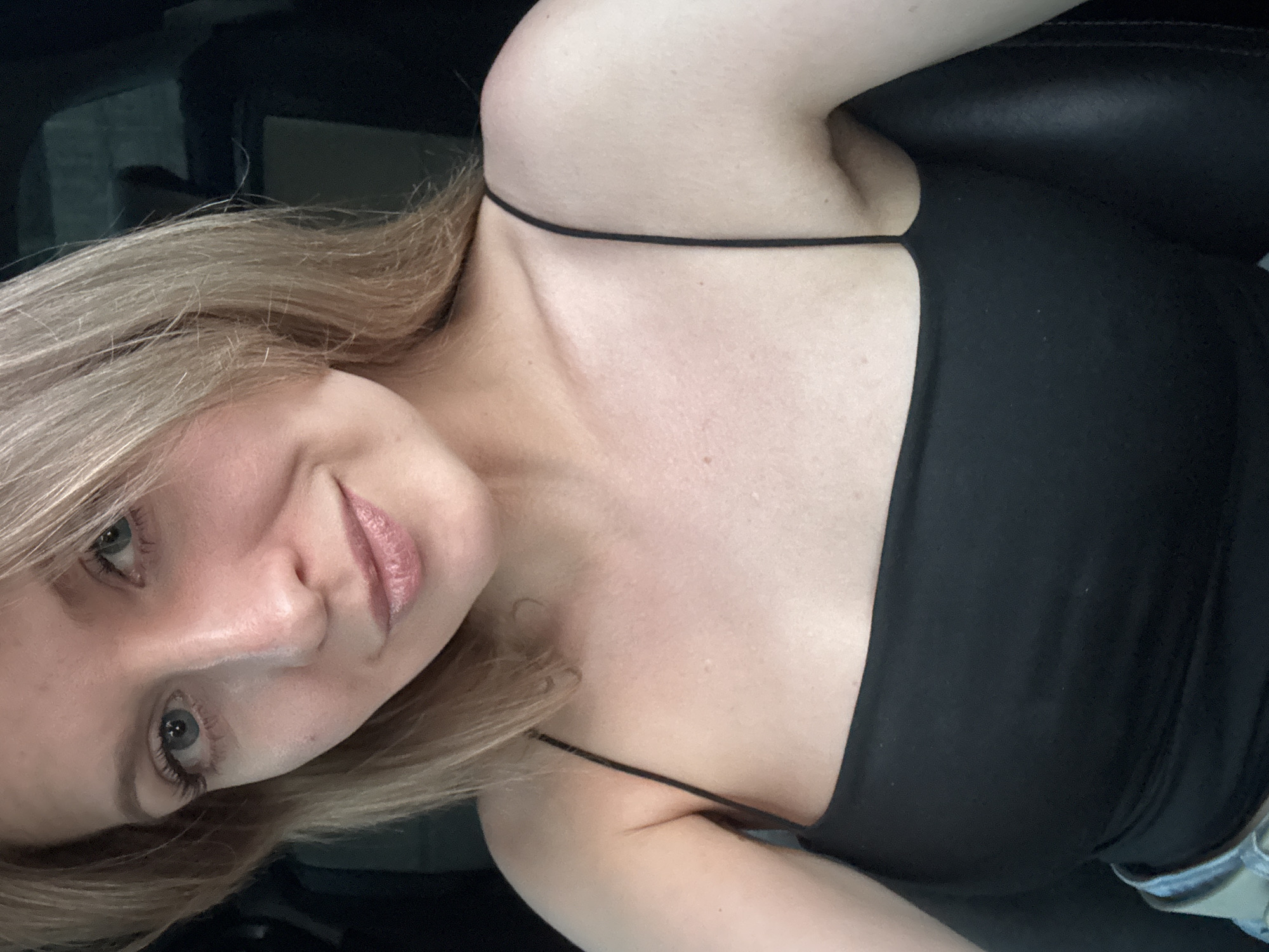 A fair-skinned young woman with light blonde hair and blue eyes is shown in a close-up, selfie-style photograph. She is wearing a black tank top with thin straps. The background appears to be the interior of a vehicle, with dark upholstery or panels visible. There is no indication of this image being taken in Izhevsk, Russia.