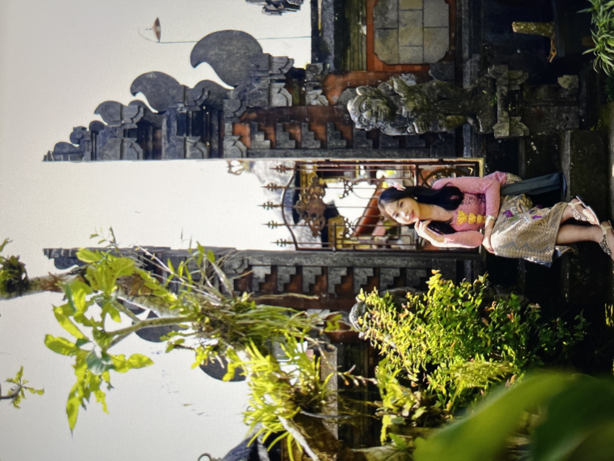 In an outdoor setting that appears to be a temple complex or traditional Indonesian courtyard, a young woman is seated on stone steps. She is wearing a traditional pink blouse with yellow embellishments and a patterned skirt. Her hair is dark and long, with a pink flower tucked behind her ear. She is looking towards the camera with a gentle smile, her right hand resting on her chin.
The backdrop features intricate stone architecture, characterized by carved details and layered stonework, consistent with Balinese or Javanese temple design. To the left, lush green foliage and vines are abundant, with some plants growing on the stonework itself. A weathered stone sculpture of a mythical creature stands to the right of the woman, adding to the cultural and historical ambiance. The sky is bright and appears overcast or hazy. The overall scene conveys a sense of cultural tradition and serene beauty. The location is identified as Cirahayu, Indonesia.