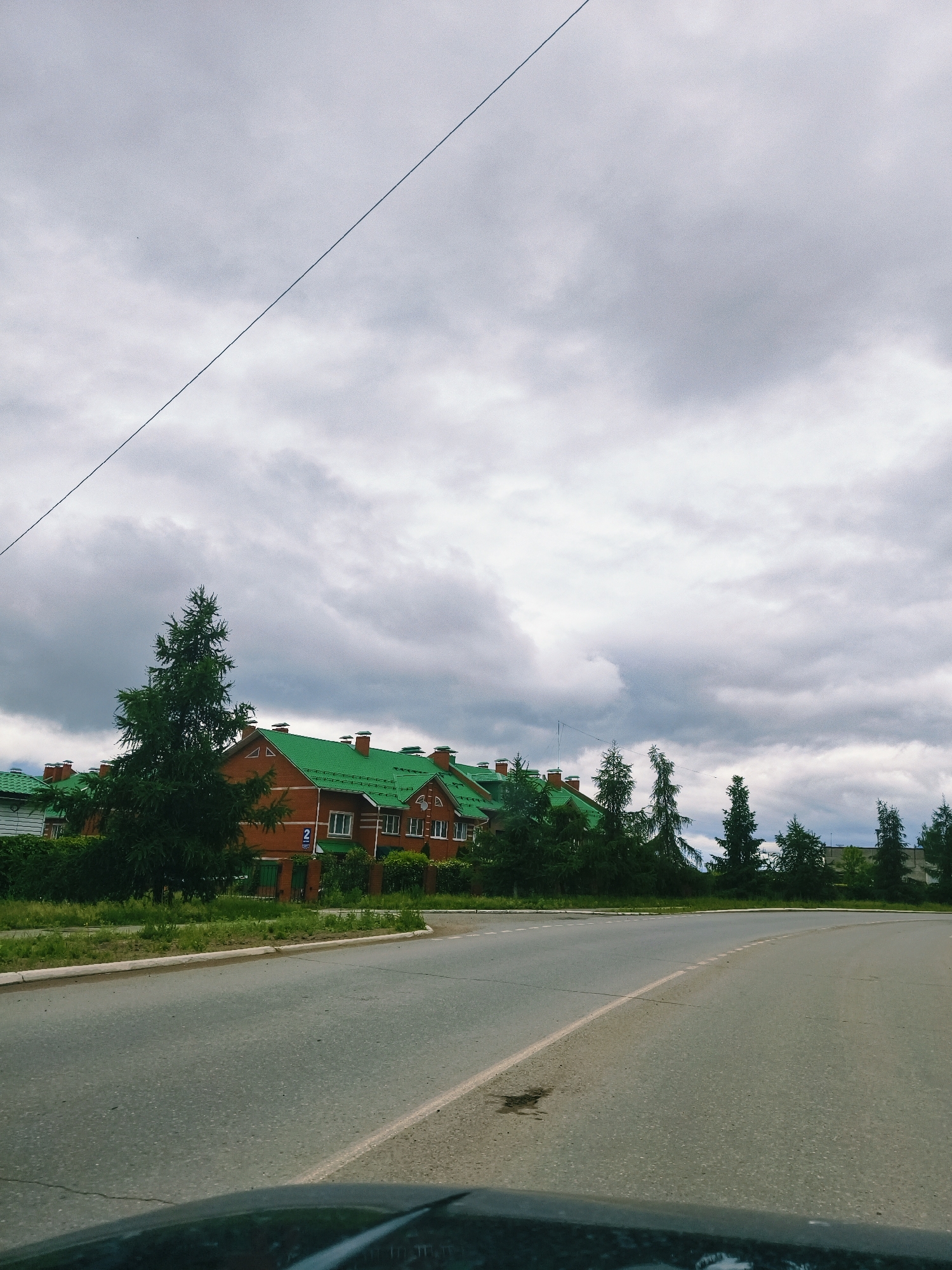 The image depicts a residential street scene under a heavily overcast sky. The viewpoint is from a vehicle, with a portion of a dark dashboard or hood visible in the lower foreground. An asphalt road, marked with a solid white line on its right edge and a broken white center line, curves gently to the right, extending from the foreground into the mid-ground. A small, dark, wet stain is present on the road surface in the left mid-ground.
To the left of the road, a concrete curb separates the asphalt from a grassy verge. Beyond the verge, a row of red-brick houses with prominent bright green pitched roofs is visible. These houses appear to be either attached or semi-detached, featuring multiple chimneys and white-framed windows. One specific house displays a blue house number sign with the numeral "2" clearly visible. Dark green coniferous trees are densely planted along the roadside, interspersed among the houses, and form a denser line behind them, extending further into the background.
The sky is filled with dense, grey clouds, indicating overcast weather conditions. A thin, dark utility line traverses the upper left quadrant of the frame diagonally, from the upper left corner downwards towards the right. No individuals or active subjects are present in the frame. The scene is located in the city of Chaikovsky, Russia.