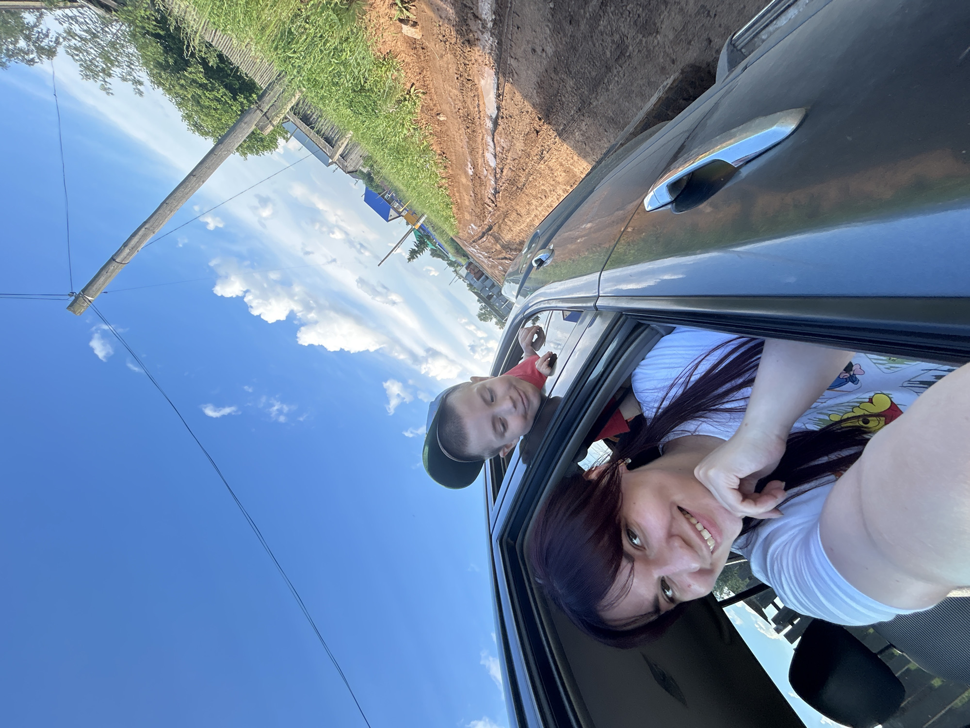 A low-angle, eye-level, outdoor, medium shot captures a woman in the front passenger seat of a vehicle and a boy leaning out of the rear window. The woman is in the foreground, smiling broadly as she takes the selfie with her right hand. She has long, dark hair and is wearing a white t-shirt with a graphic print. The boy, wearing a red shirt and a green baseball cap, is in the background, looking towards the woman with a slight smile.
The vehicle's interior is visible, including the black headrest of the driver's seat. Outside the car, a dirt road with muddy puddles stretches into the distance. On the right side of the road, a dense green grassy embankment is present, along with some wooden fences and buildings. In the background, a tall wooden utility pole with wires stretches across the frame, supporting electrical or communication lines. The sky is bright blue with scattered white clouds. The scene appears to be in a rural area, possibly on the outskirts of the city of Fokino, Russia, given the context of the prompt.