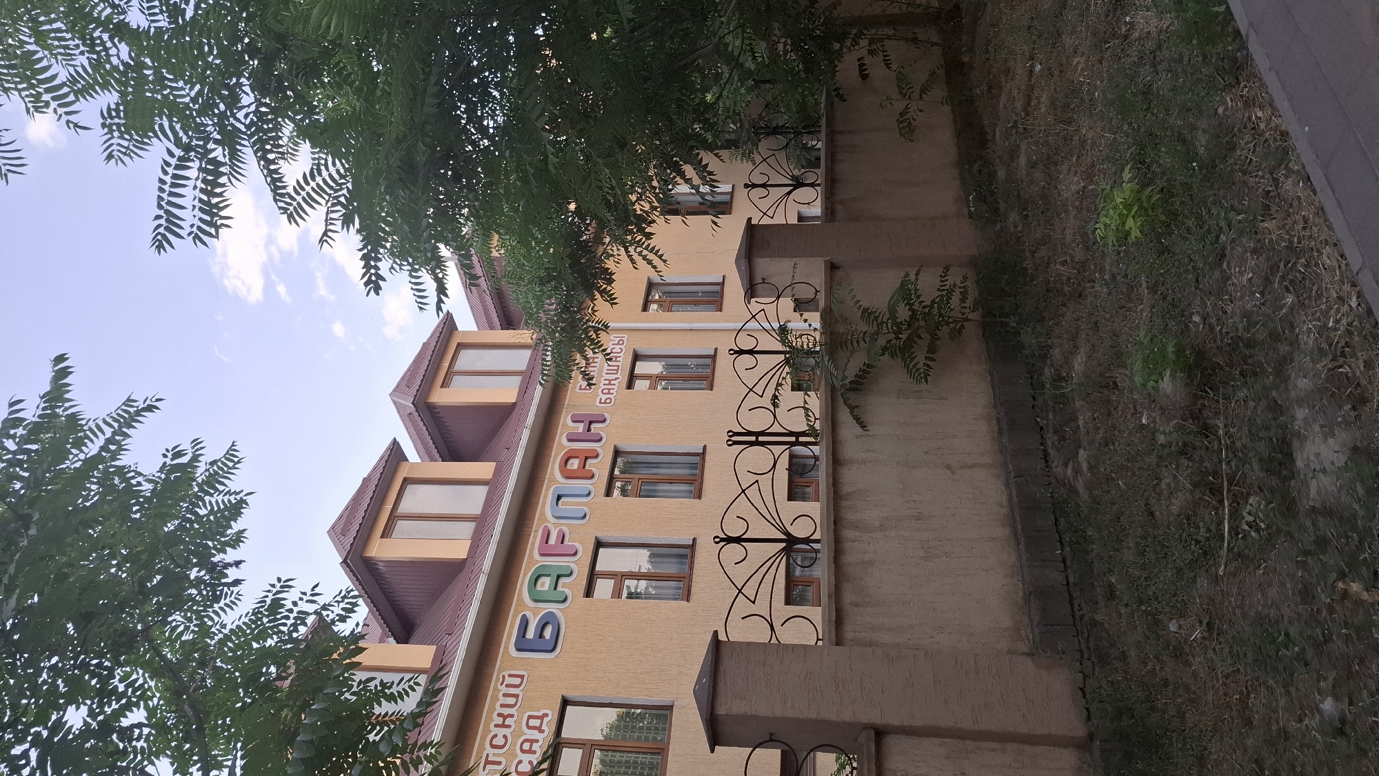 The image shows a light orange building with a red roof and dormer windows. Above the windows, there are large, colorful letters spelling out "БАГЛАН БАКШАСЫ" and some Cyrillic text to the left, "ДТСКИЙ САД". This translates to "Baglan Kindergarten". The building is partially obscured by green trees. A beige concrete fence with decorative black metalwork runs along the front of the property, separating it from a grassy area with a paved walkway. The scene appears to be outdoors in daylight, with a cloudy sky visible. The location is specified as Shymkent, Kazakhstan.