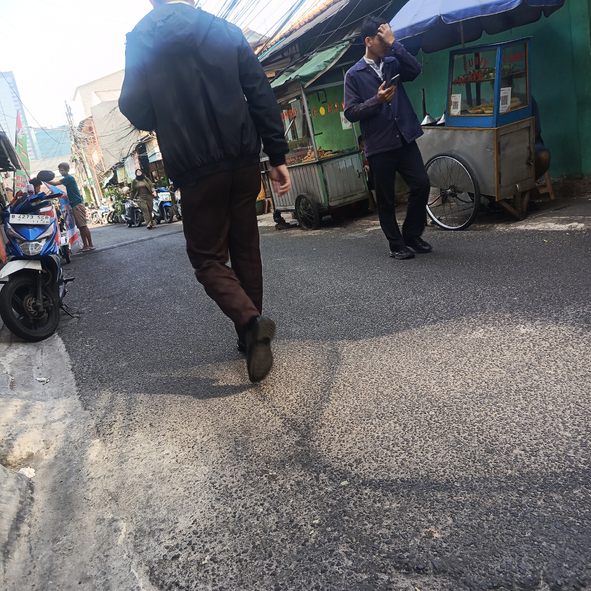 The image shows a street scene in South Jakarta, Indonesia, likely in an urban area with dense housing or commercial activity. In the foreground, a person wearing a dark jacket and brown pants walks away from the camera. To the right, another person in a dark purple jacket and black pants stands holding a mobile phone, with a street food cart beside them. The cart appears to be selling fried food, with a glass display showing some items. Several motorcycles are parked on the street, and a blue and white motorcycle is prominently featured on the left side of the image. The street is paved with asphalt and shows signs of wear. Buildings, some with awnings and signs in Indonesian, are visible in the background, along with a tangle of overhead electrical wires, typical of urban environments in Indonesia. The scene appears to be illuminated by daylight, suggesting it was taken during the day.