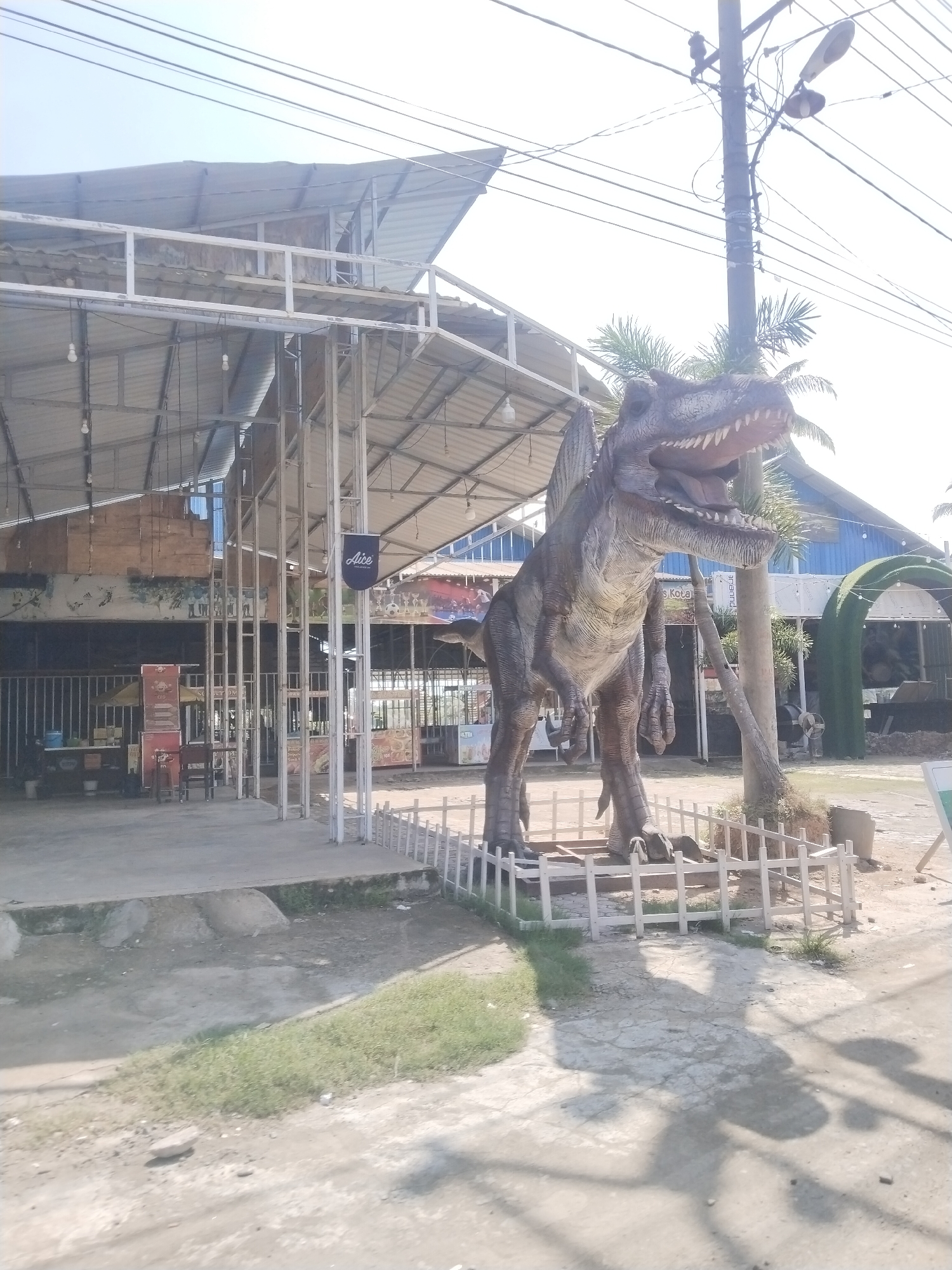 A large, realistic dinosaur statue stands prominently in the foreground, its mouth open in a roar. The statue is positioned on a concrete base surrounded by a white picket fence. Behind the dinosaur, a building with a corrugated metal roof and exposed metal framing is visible. A blue building with a curved green archway is also present in the background, suggesting an outdoor venue or park. Utility poles with numerous wires crisscross the sky. The ground is a mixture of cracked concrete and patches of dry grass. The lighting suggests it is daytime with clear skies and bright sunlight, casting distinct shadows of the dinosaur and the surrounding structures. The overall scene appears to be an outdoor attraction or theme park, possibly in a commercial area, given the presence of signage and the architectural style of the buildings. The location is Lhokseumawe, Indonesia.
