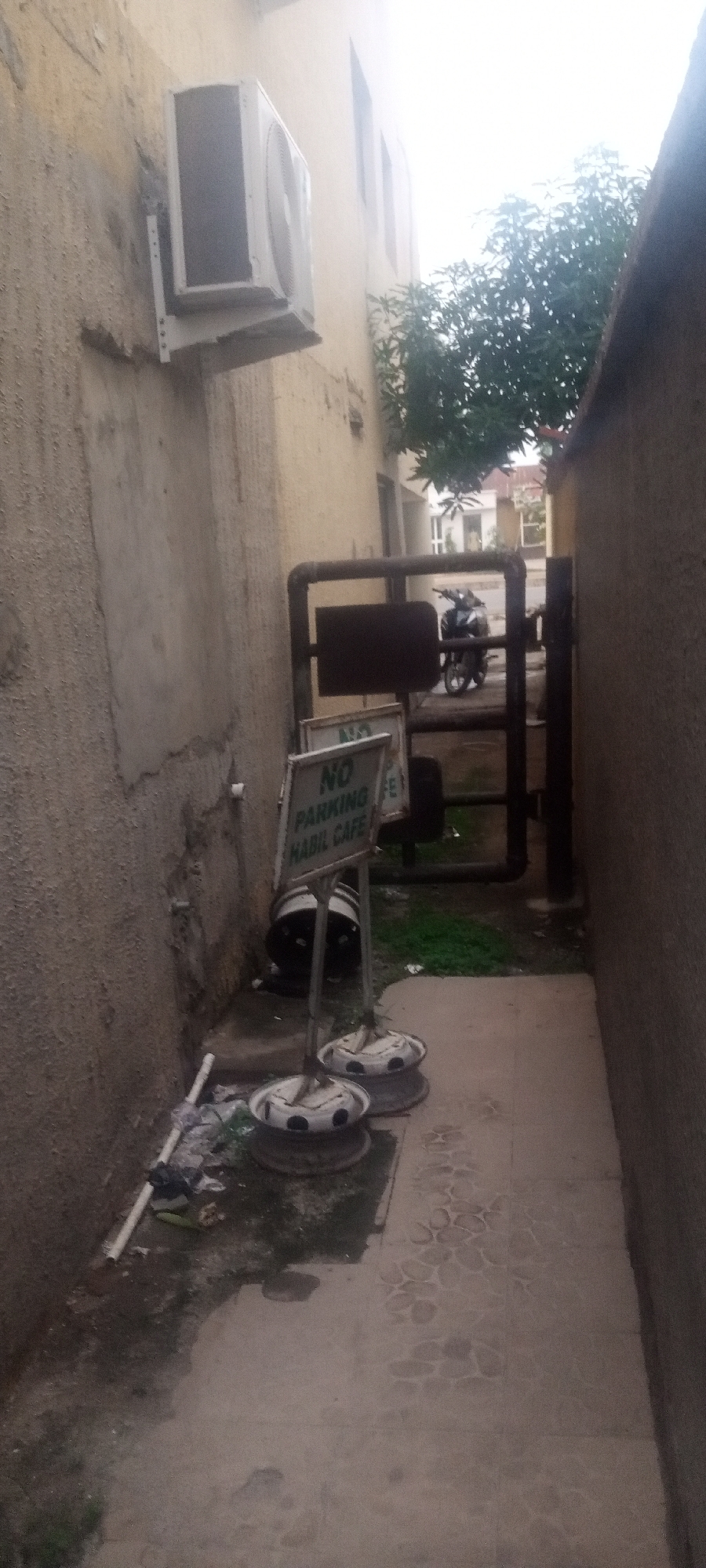 The image depicts a narrow passageway in an urban environment. A "No Parking HABIL CAFE" sign is present, suggesting the location is associated with that establishment. Two car wheels appear to act as bases for the sign. A metal gate or barrier is visible further down the passageway, behind which a motorcycle is parked. The passageway is bordered by the exterior walls of buildings, and an air conditioning unit is mounted on one of the walls. Debris and litter are scattered along the base of the wall on the left side. The ground is tiled. The presence of the "Habil Cafe" sign suggests the location is Kabala, Nigeria.