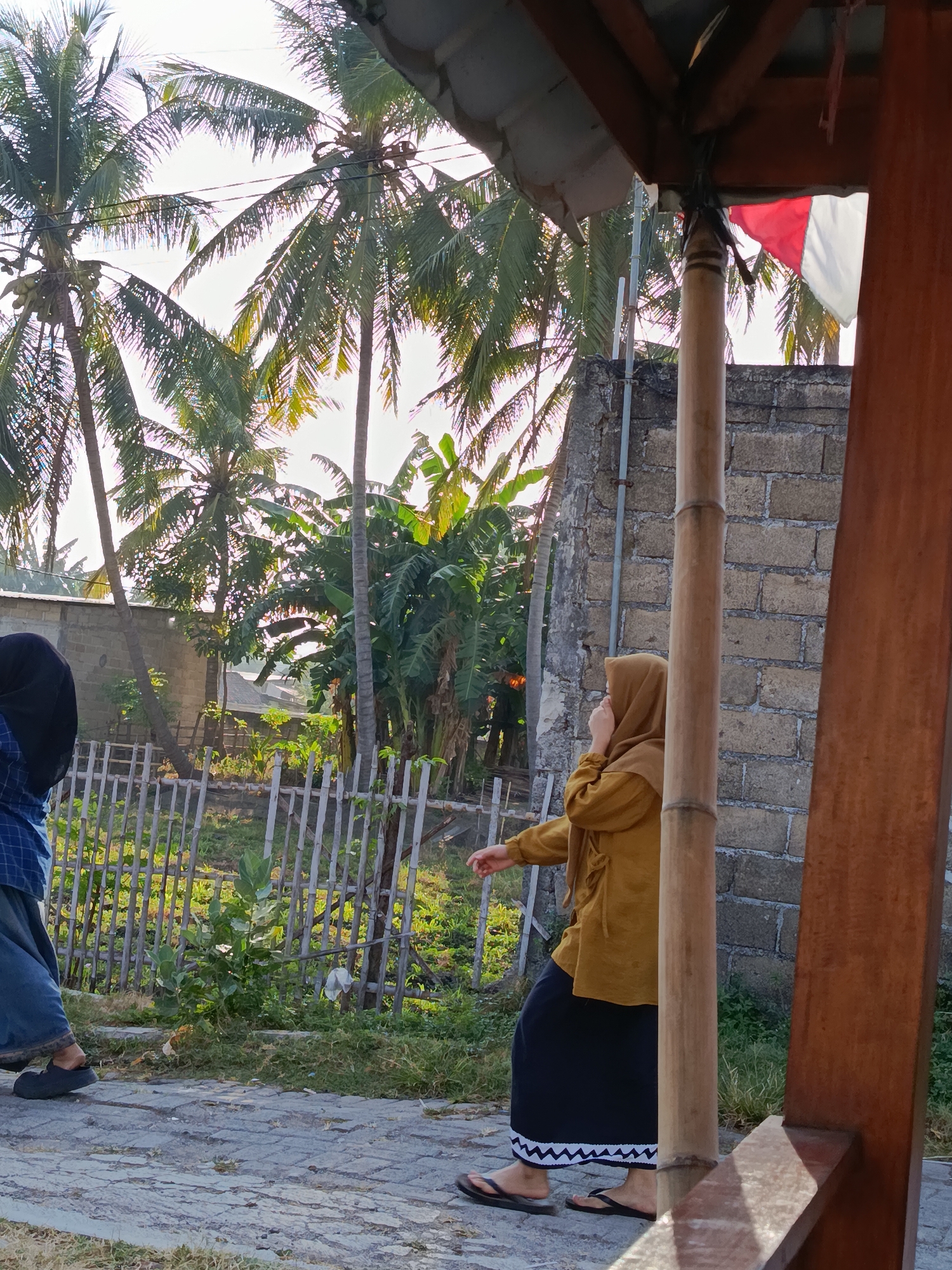 The image depicts an outdoor scene in Ramban Wetan, Indonesia, with a focus on natural elements and a building structure. In the foreground, a pathway paved with stone blocks leads towards a wooden veranda or shelter. A woman wearing a mustard-colored hijab and a long black skirt with a white zig-zag pattern is standing on the pathway, with her right hand raised to her face. To her left, another person, partially visible and wearing a blue plaid shirt and blue pants, is also on the pathway.
Behind the pathway, a rustic bamboo fence encloses a garden area with lush green foliage and some vegetation. Beyond the fence, a brick wall stands, and further in the background, several tall coconut trees and banana trees are visible against a bright, overcast sky. A small Indonesian flag, red and white, is attached to the wooden structure in the upper right quadrant of the image. The overall impression is a tranquil, tropical setting.