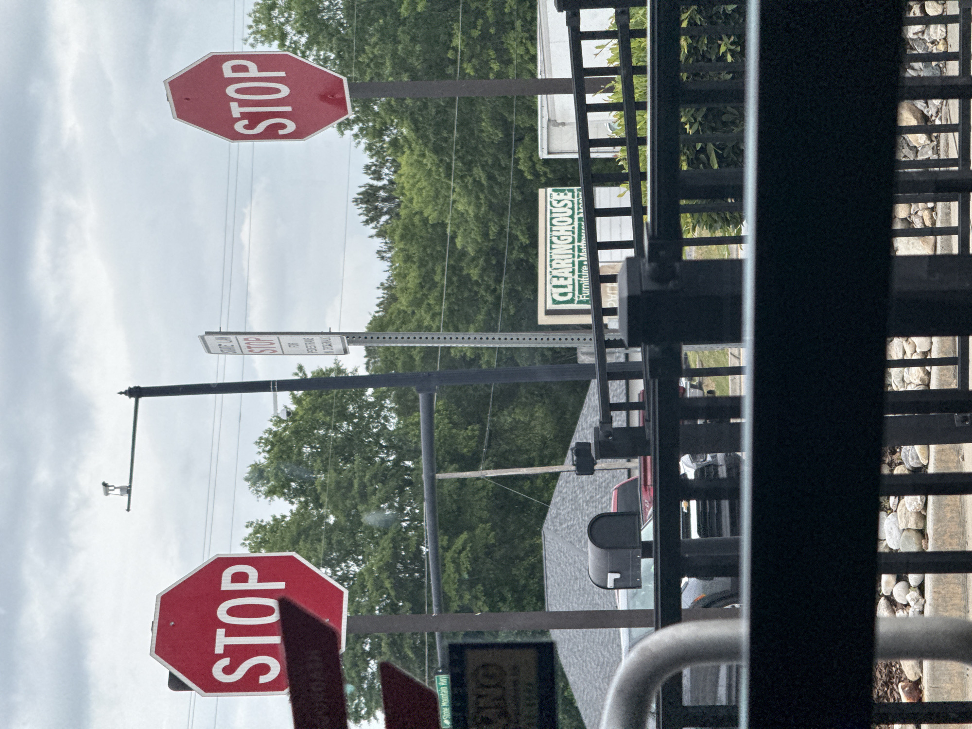 The image shows a scene in Stone Mountain, Georgia, United States. There are two stop signs and a sign that says "Clearinghouse Furniture, Mattress, and More". A street sign for Stone Mountain Hwy is visible. There is a black metal fence in the foreground. The weather is overcast. There are trees in the background. A utility pole and wires are visible. Cars are parked on the street.