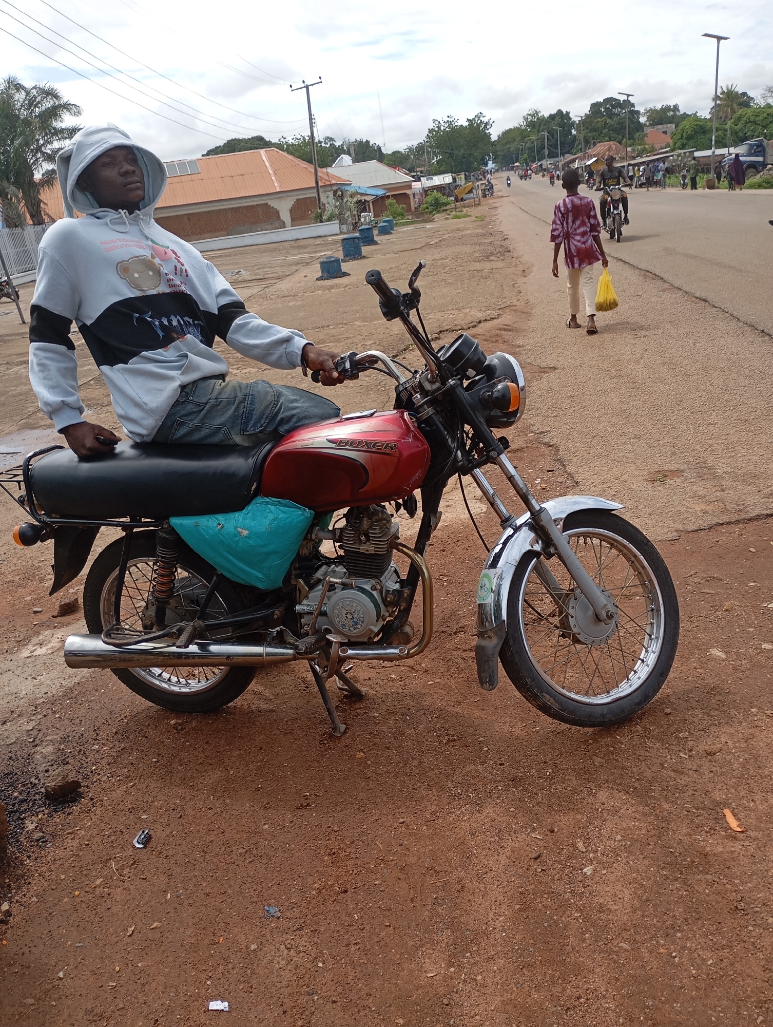 In Shendam, Nigeria, a young man sits on a red motorcycle on a dirt road. He wears a light grey hoodie with black and white accents and blue jeans. The motorcycle has a black seat and a turquoise bag tied to its side. In the background, a street stretches into the distance, lined with buildings and trees. Other people and vehicles are visible on the street, and a few utility poles stand alongside it. The sky is overcast.
