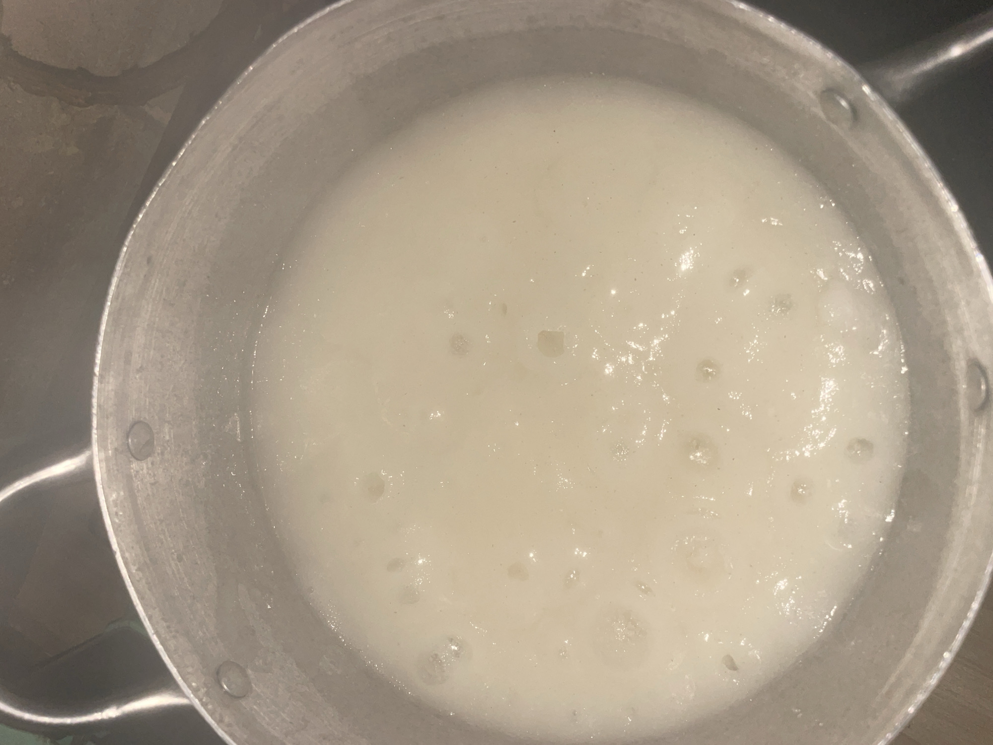A top-down, close-up shot shows a pot of what appears to be a starchy, white liquid bubbling gently. The pot itself is a metallic, possibly aluminum material, with a slightly worn texture. The liquid has a creamy, opaque appearance, with small bubbles breaking the surface in various locations, suggesting it is being heated. The rim of the pot is visible, with rivets securing the handles. The background is out of focus and dark, hinting at an indoor setting. There are no people or animals visible in the frame. The image does not contain any elements that would allow for identification of a specific city like Lokoja, Nigeria.
