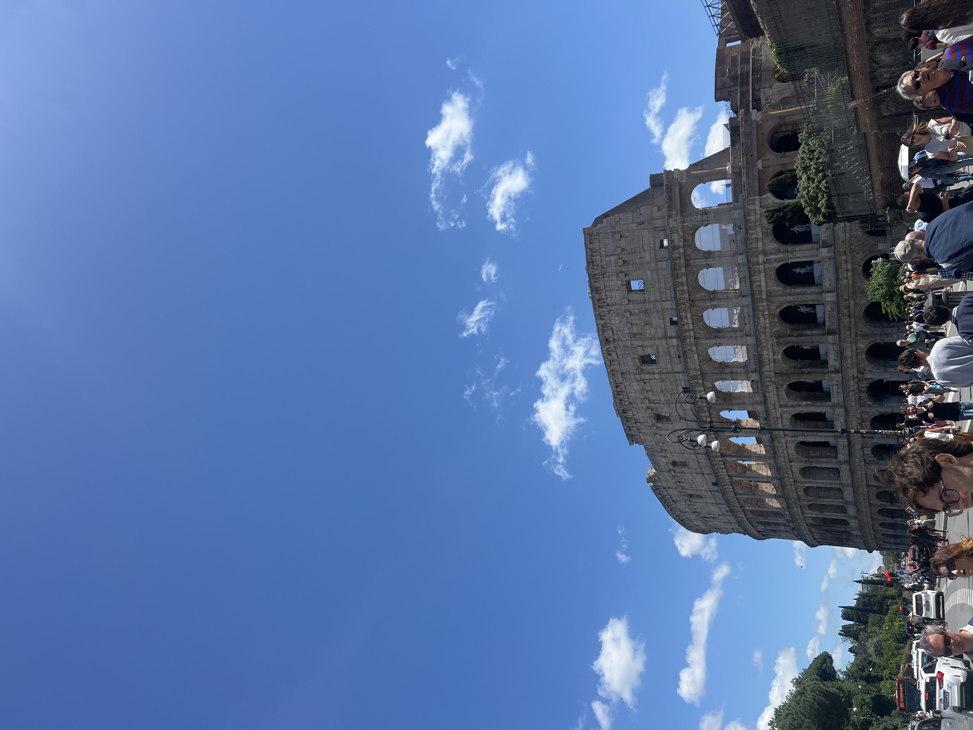 The image shows the Colosseum in Rome, Italy, under a blue sky with scattered white clouds. The ancient amphitheater is partially visible, showcasing its tiered structure and arched openings. A crowd of people is present, suggesting the site is a popular tourist destination. Trees and vehicles are visible in the lower part of the image, indicating the urban setting. The orientation of the photo is rotated, showing the sky occupying a large portion of the frame.