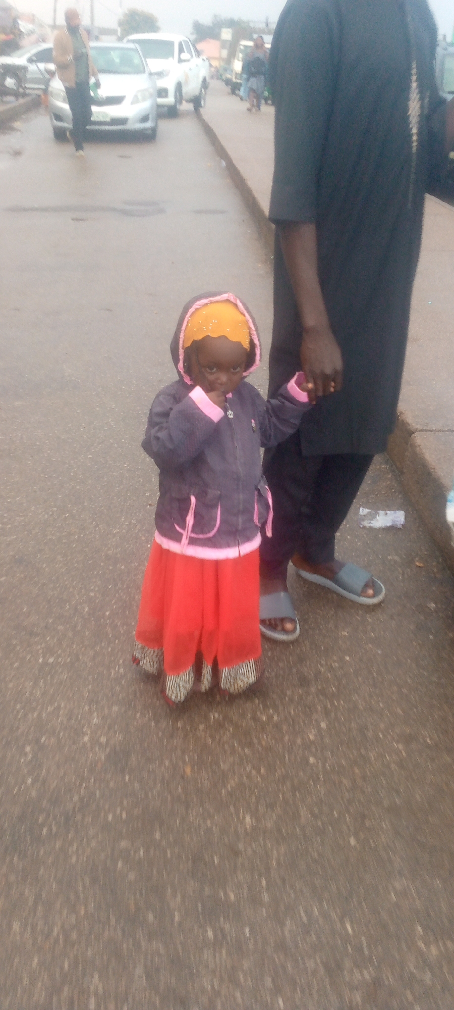 A young child wearing a yellow headscarf, a dark jacket with pink trim, and a bright red skirt is standing on an asphalt road, holding hands with an adult whose lower body is visible, wearing dark clothing and gray sandals. The child is looking at the camera and has their fingers in their mouth. In the background, several cars and a person walking are visible on the road, suggesting a street or highway environment. The ground appears wet, indicating recent rain. The lighting is overcast.